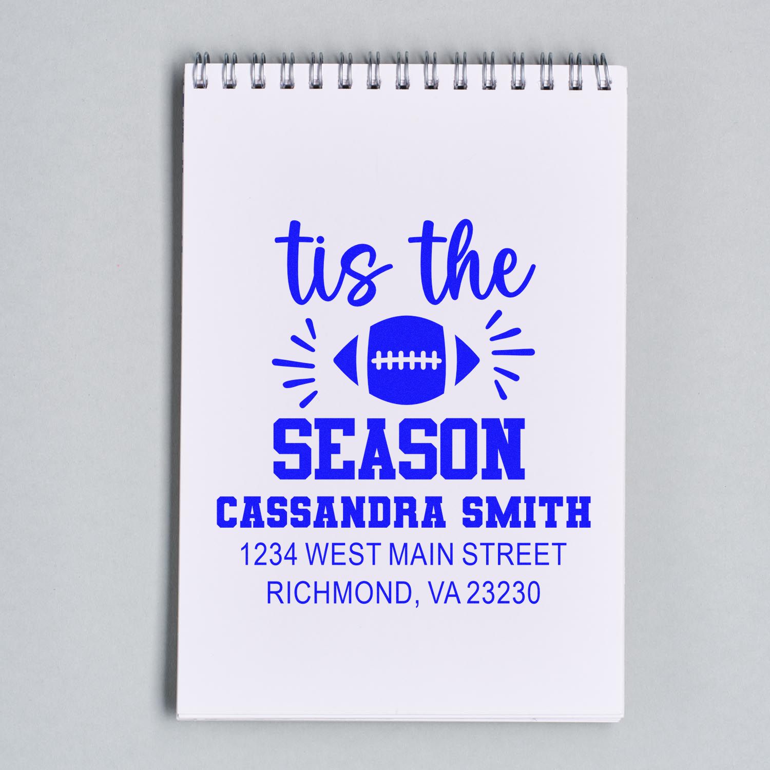 Custom rubber stamp mockup with tis the season football design, name, and address on a notebook page.