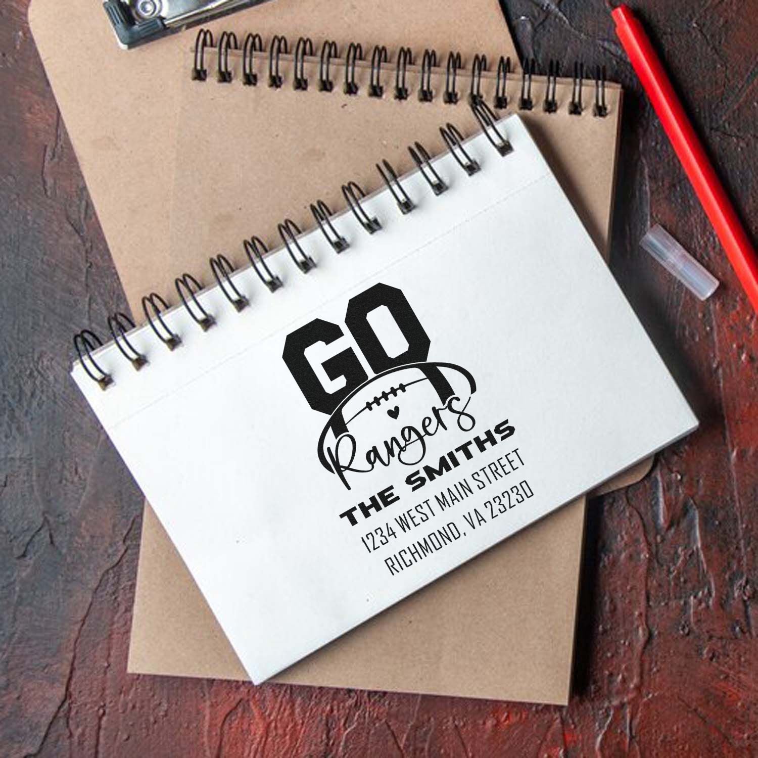 Custom rubber stamp impression on notebook, featuring a football logo, family name, and address for personalized stationery.