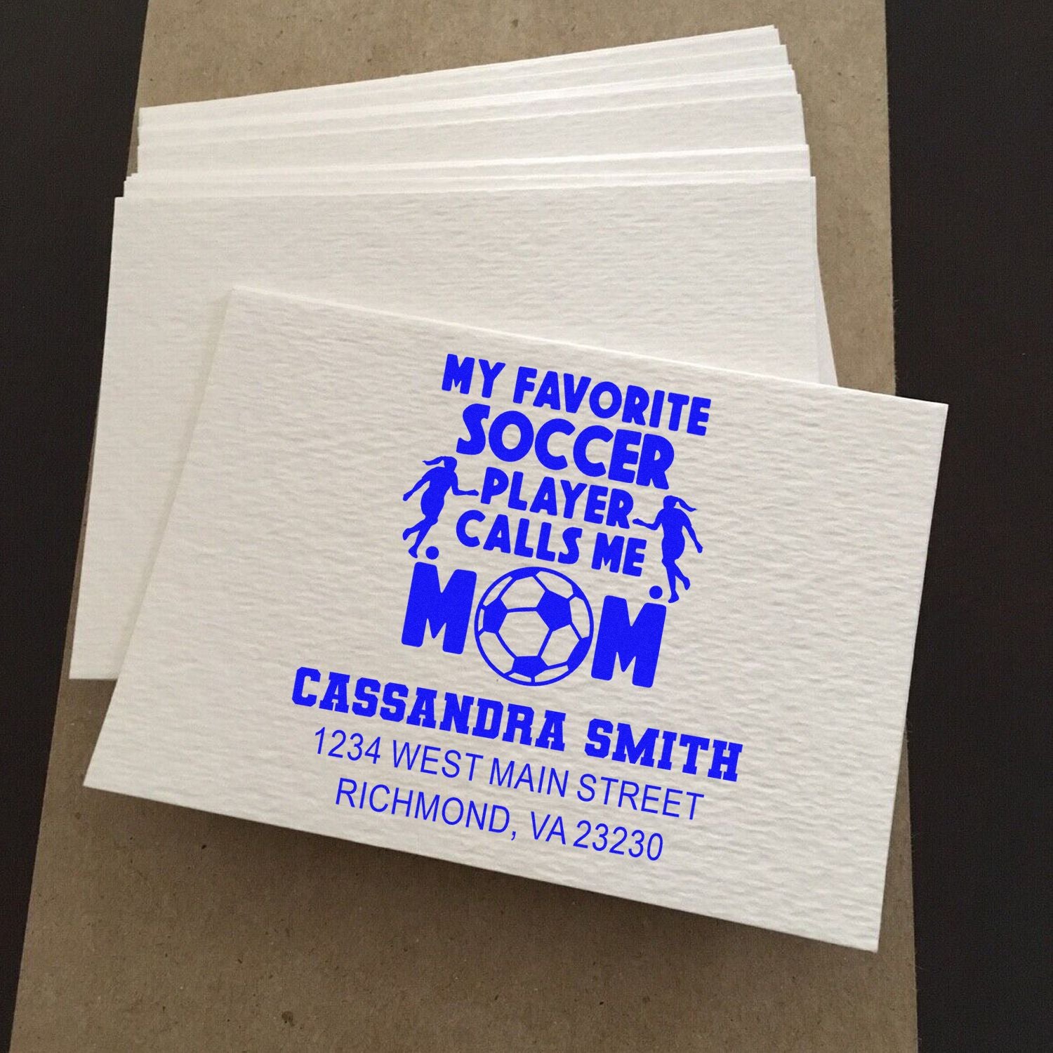 Custom rubber stamp on white napkins featuring a blue My Favorite Soccer Player Calls Me Mom design with name and address.