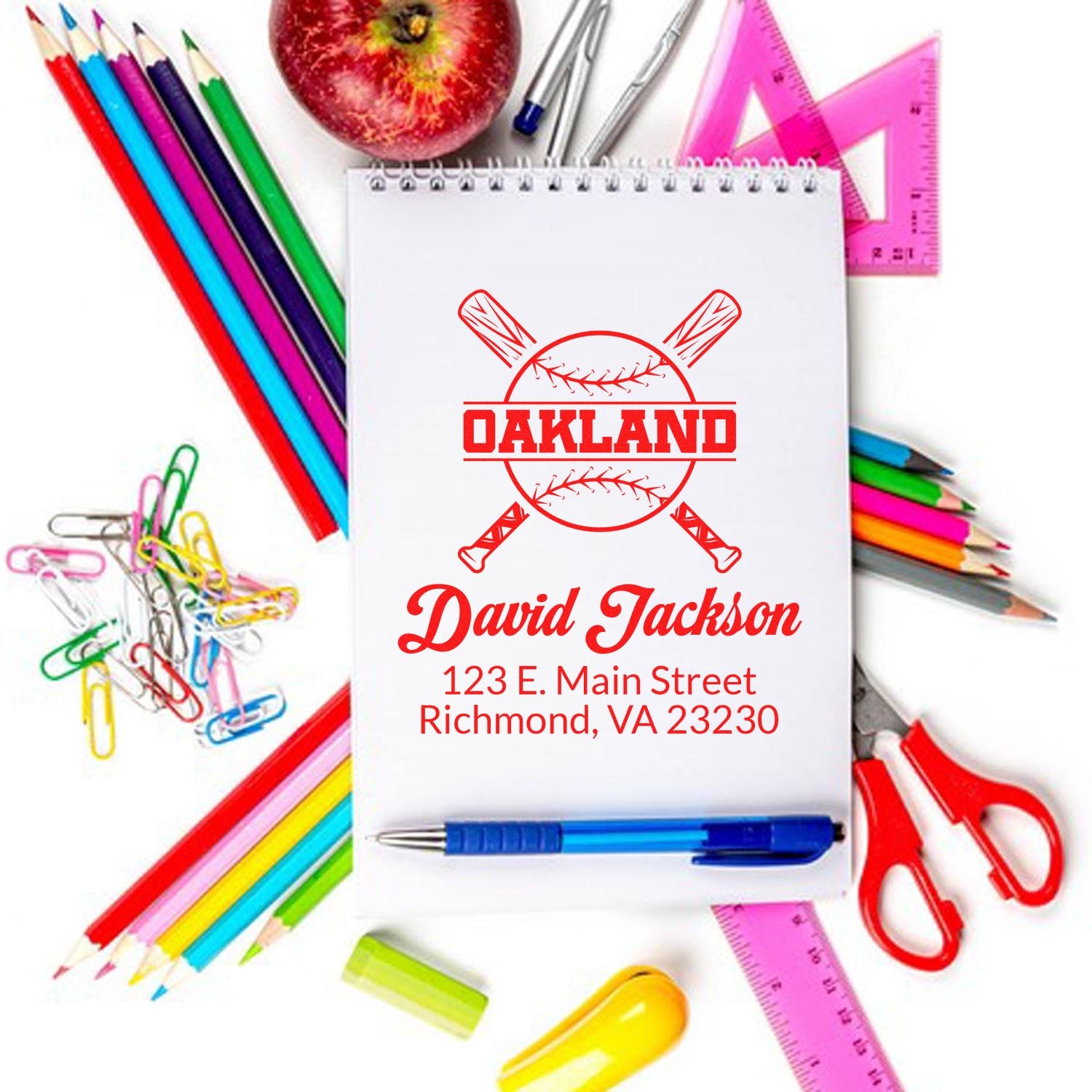 Personalized baseball rubber stamp with name and address, displayed as a red custom imprint on a notebook with school supplies.