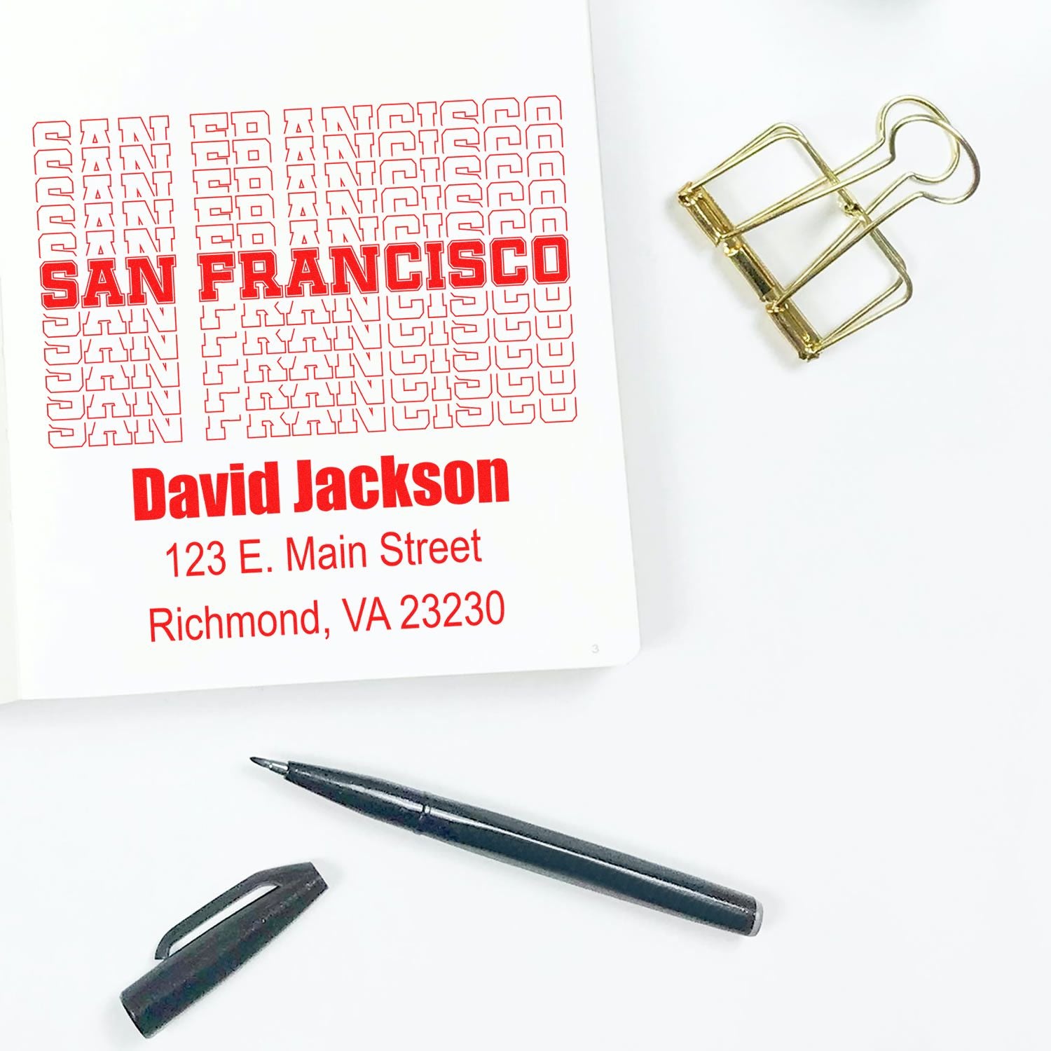 Custom rubber stamp proof with personalized San Francisco name and address design, shown with pen and binder clip.