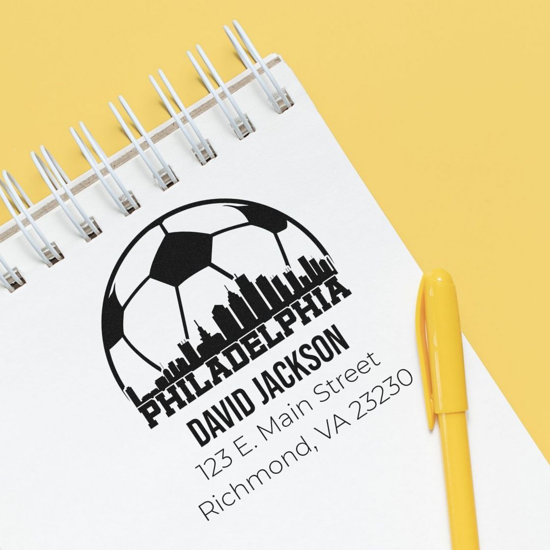 Custom Philadelphia rubber stamp with soccer ball skyline logo, business name, and mailing address on a notebook.