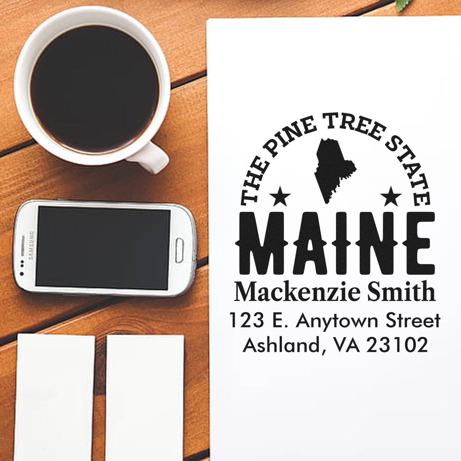 Custom Maine address rubber stamp mockup with name, street address, and state outline on paper beside coffee and phone.