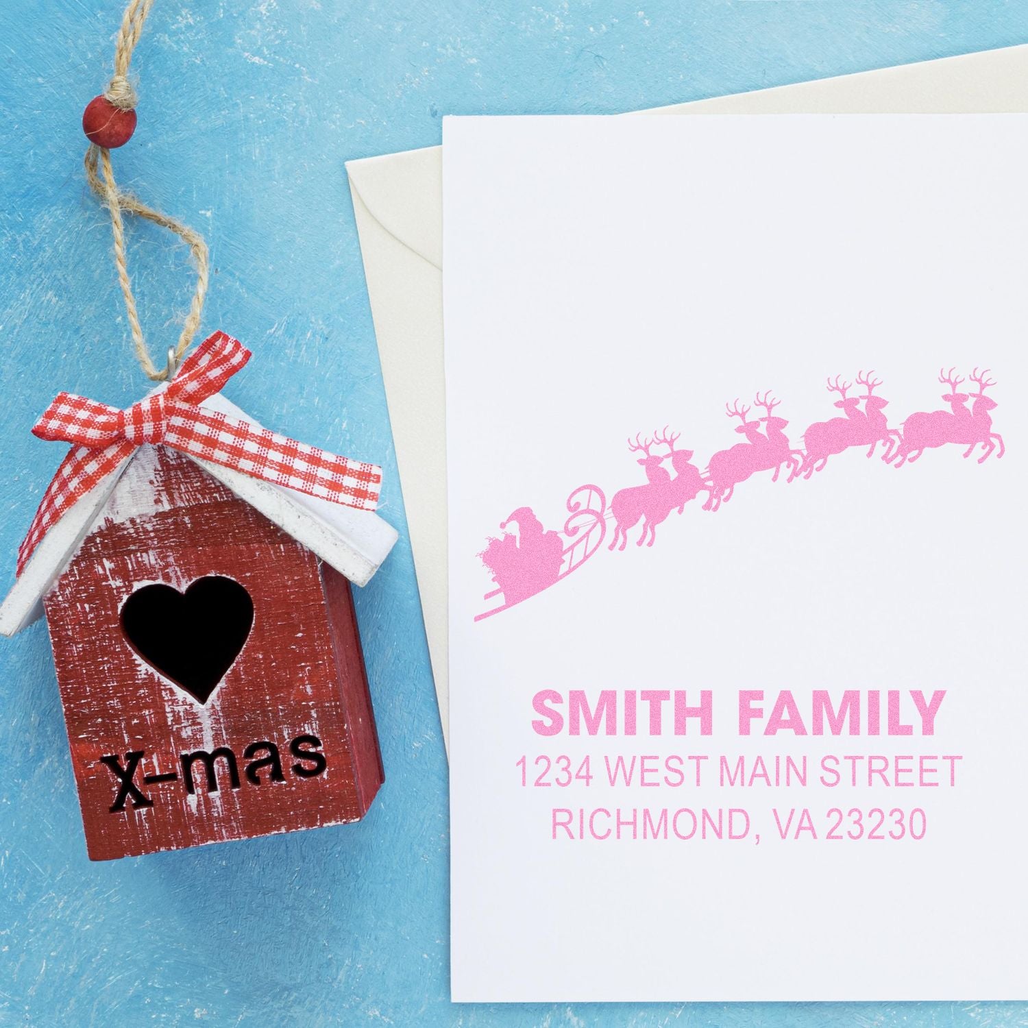 Custom Christmas rubber stamp with pink Santa sleigh return address on holiday card beside X-mas birdhouse ornament.