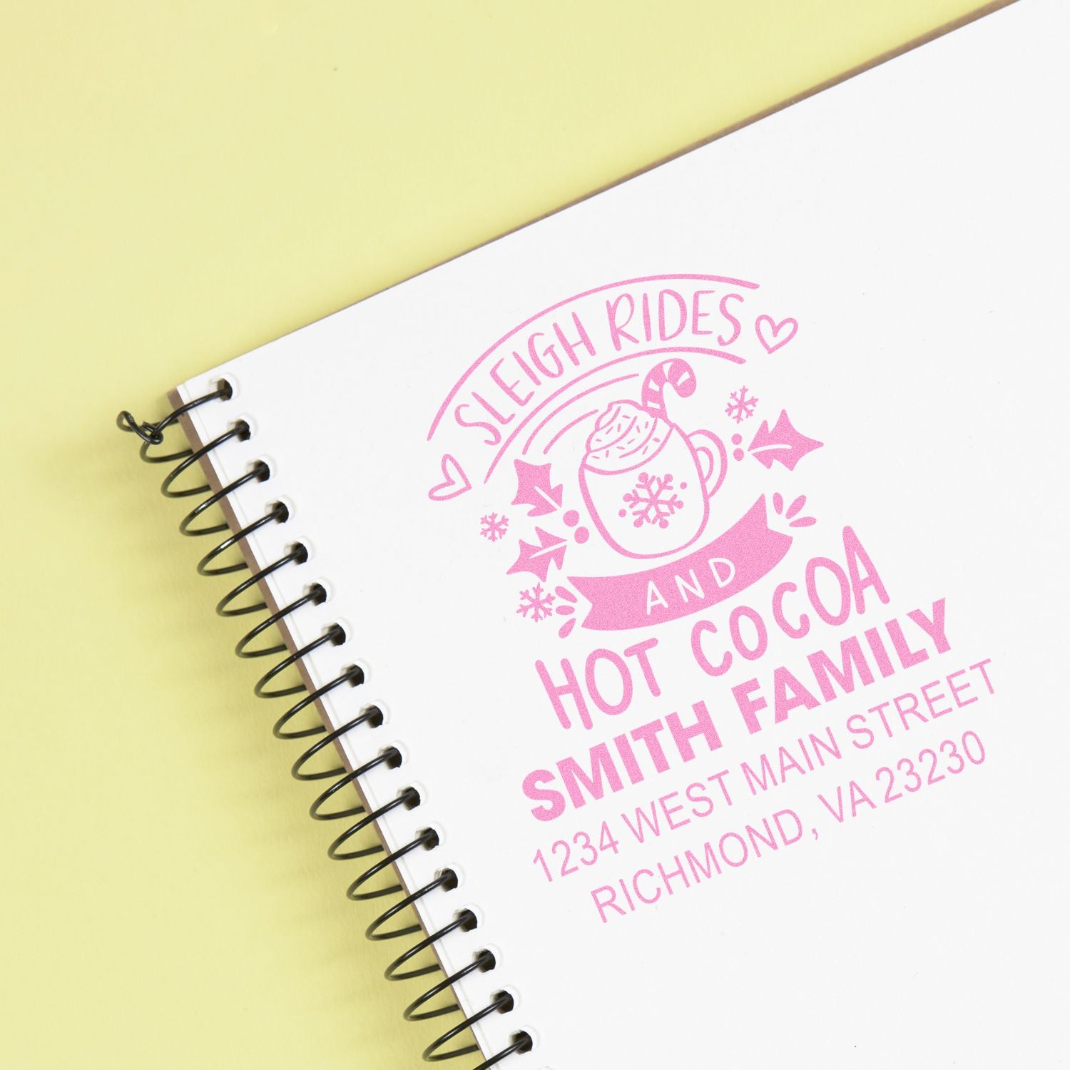 Custom pink rubber stamp on a spiral notebook for holiday branding, reading Sleigh Rides and Hot Cocoa with Family.