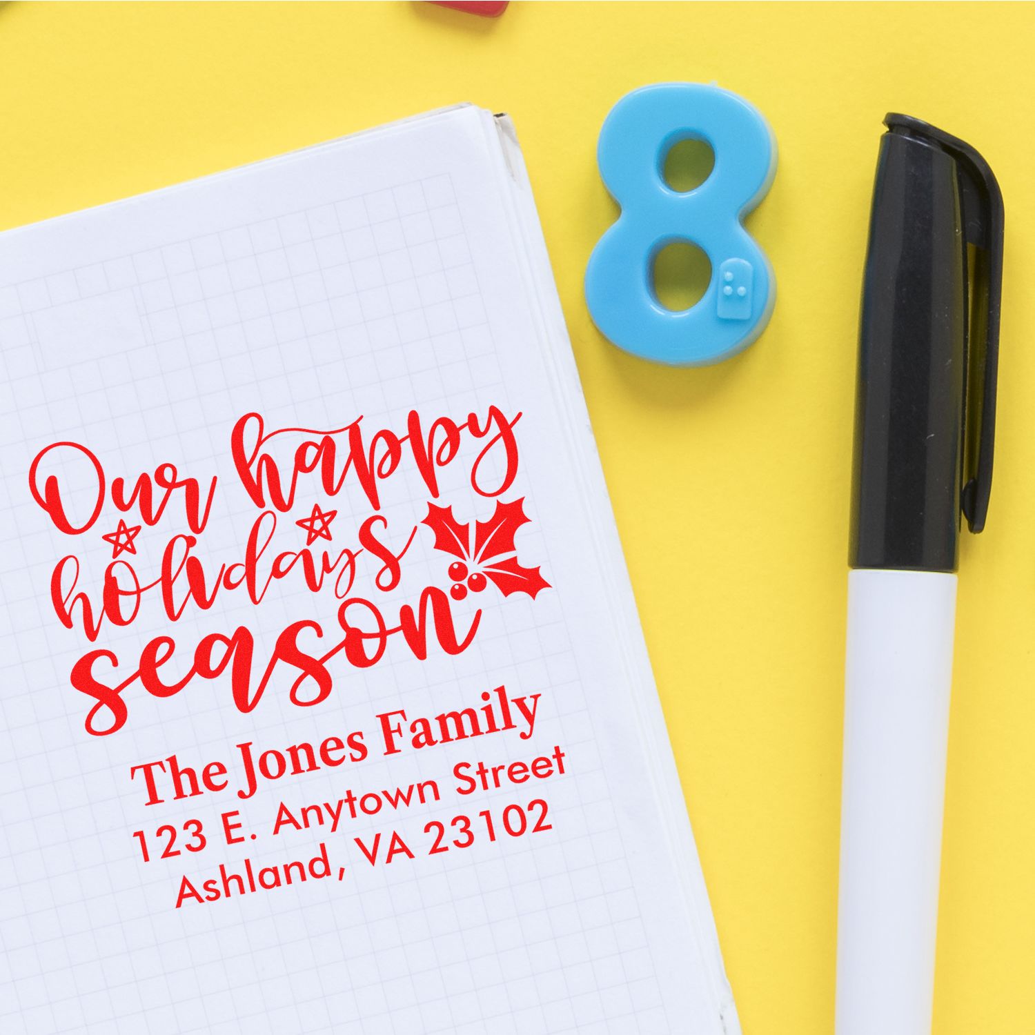 Custom holiday return address rubber stamp with festive script and family address on notebook, beside pen and number 8.