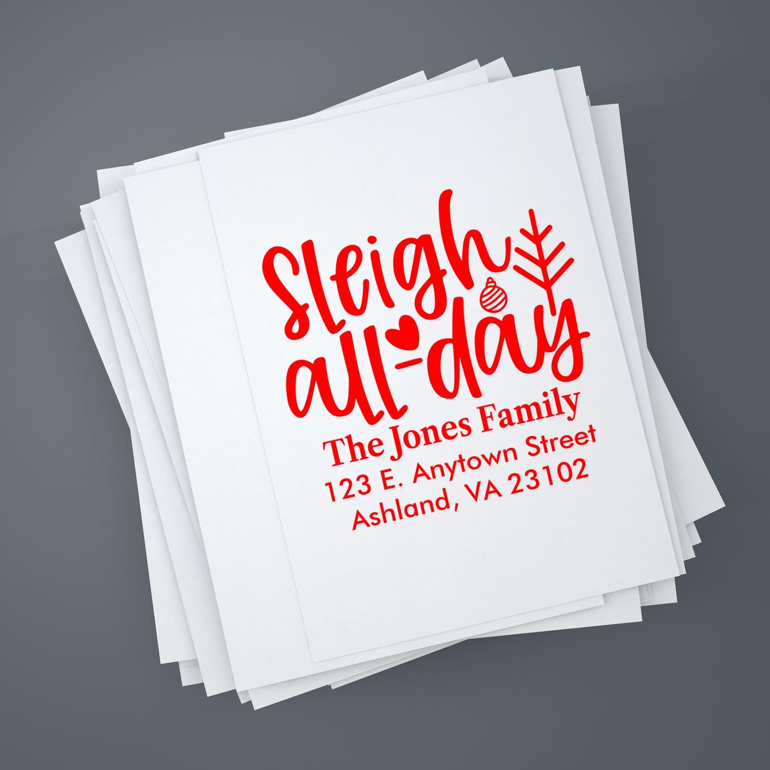 Custom holiday rubber stamp reading Sleigh All Day with family name and address on stacked paper for Christmas mailings