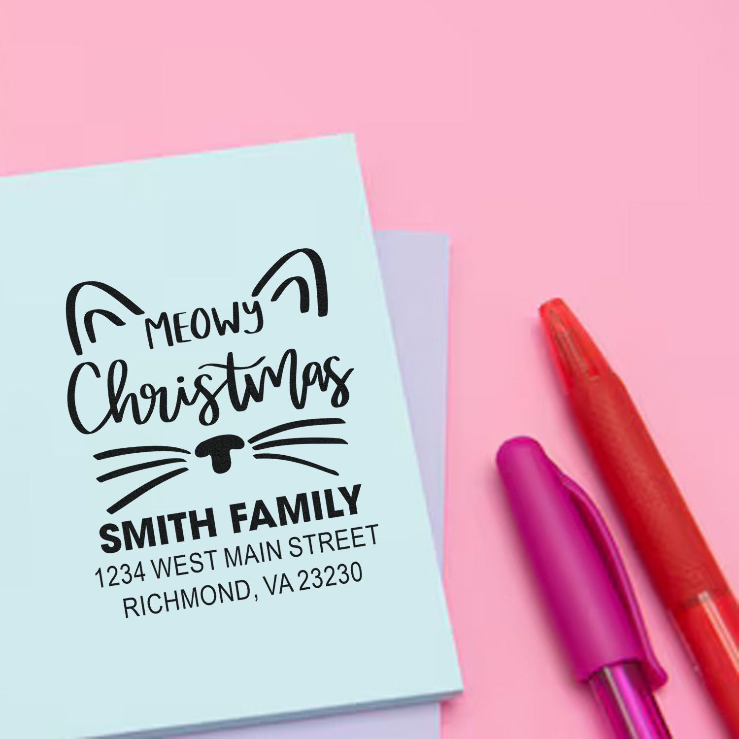 Custom cat Christmas rubber stamp with family name and return address, shown with pens for festive holiday card and envelope labeling.