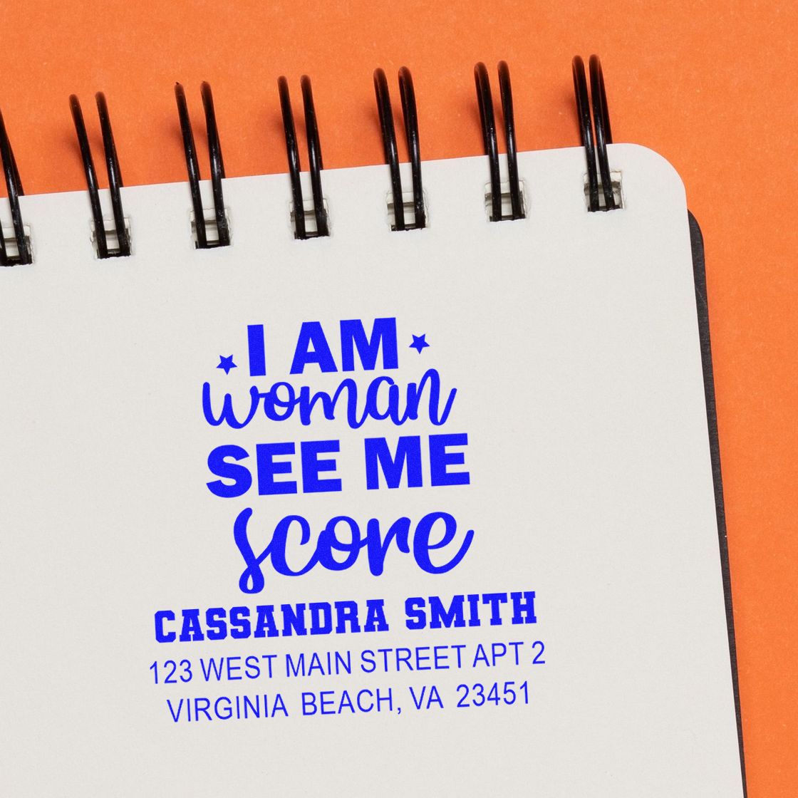 Custom rubber stamp impression with I am woman, see me score slogan, name, and address on notebook page.
