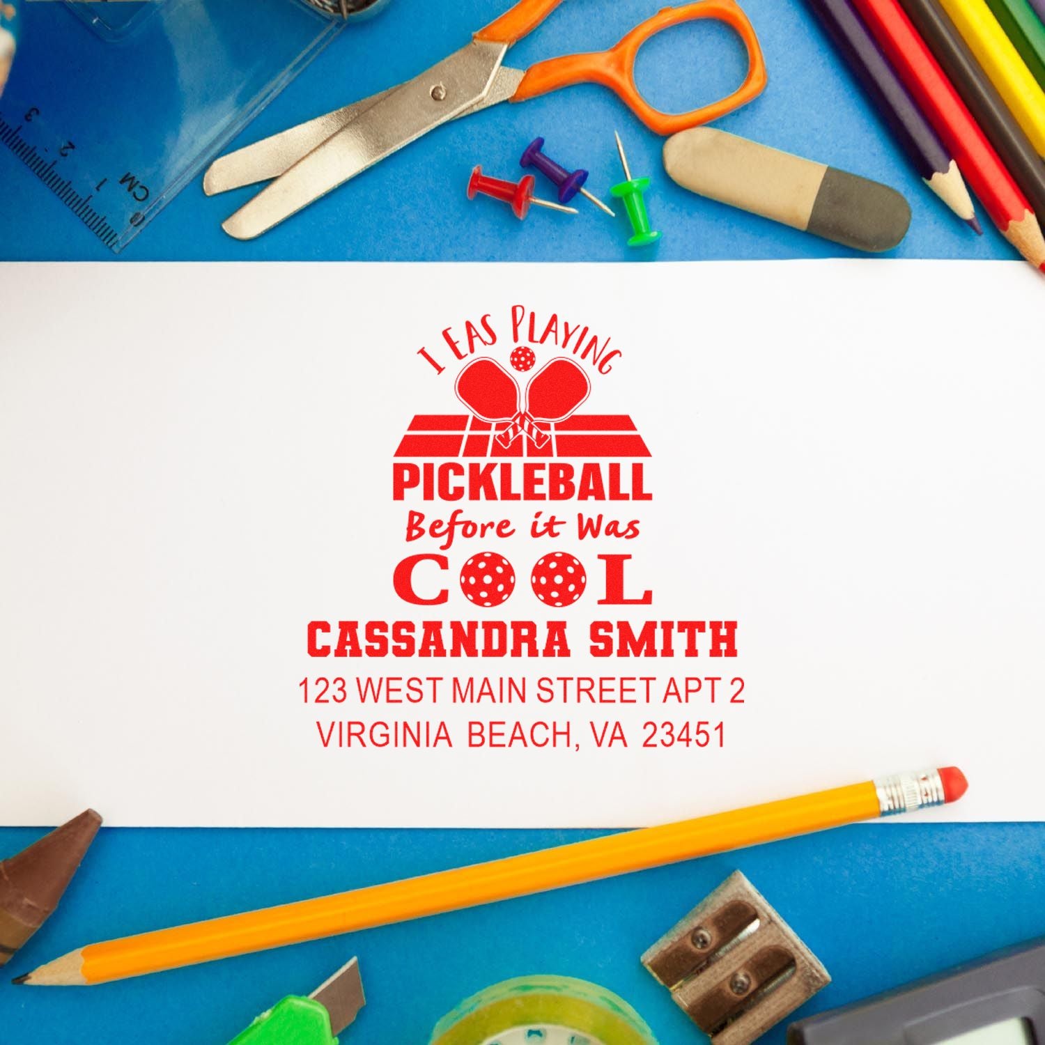 Personalized pickleball rubber stamp with name and address, stamped on paper beside school supplies and art tools.