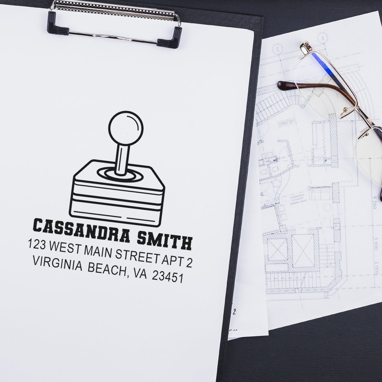 Custom personalized rubber stamp imprint with name and address on a clipboard beside architectural plans and eyeglasses.