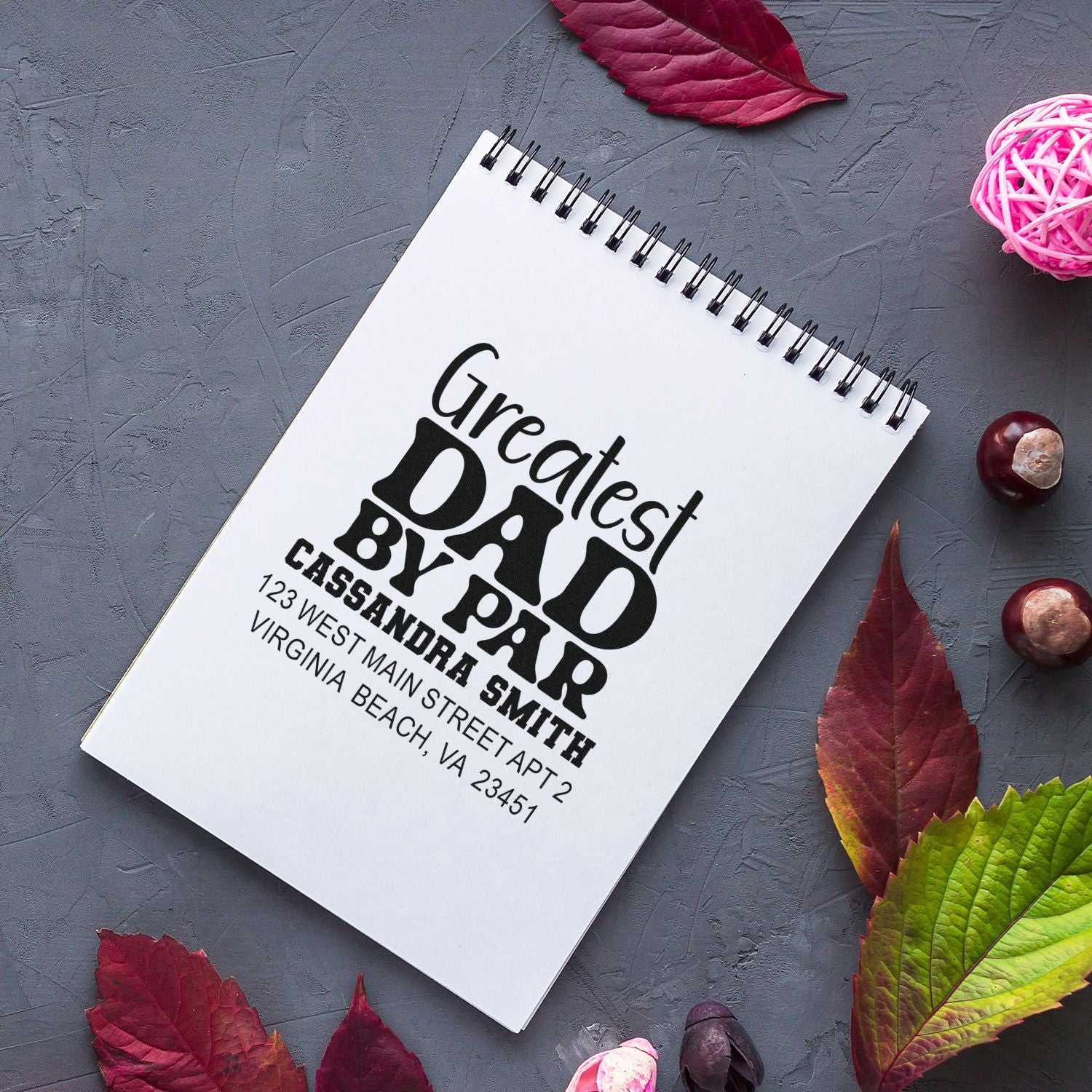 Custom rubber address stamp for a Greatest Dad gift, showing personalized family name and Virginia Beach address on a notebook.
