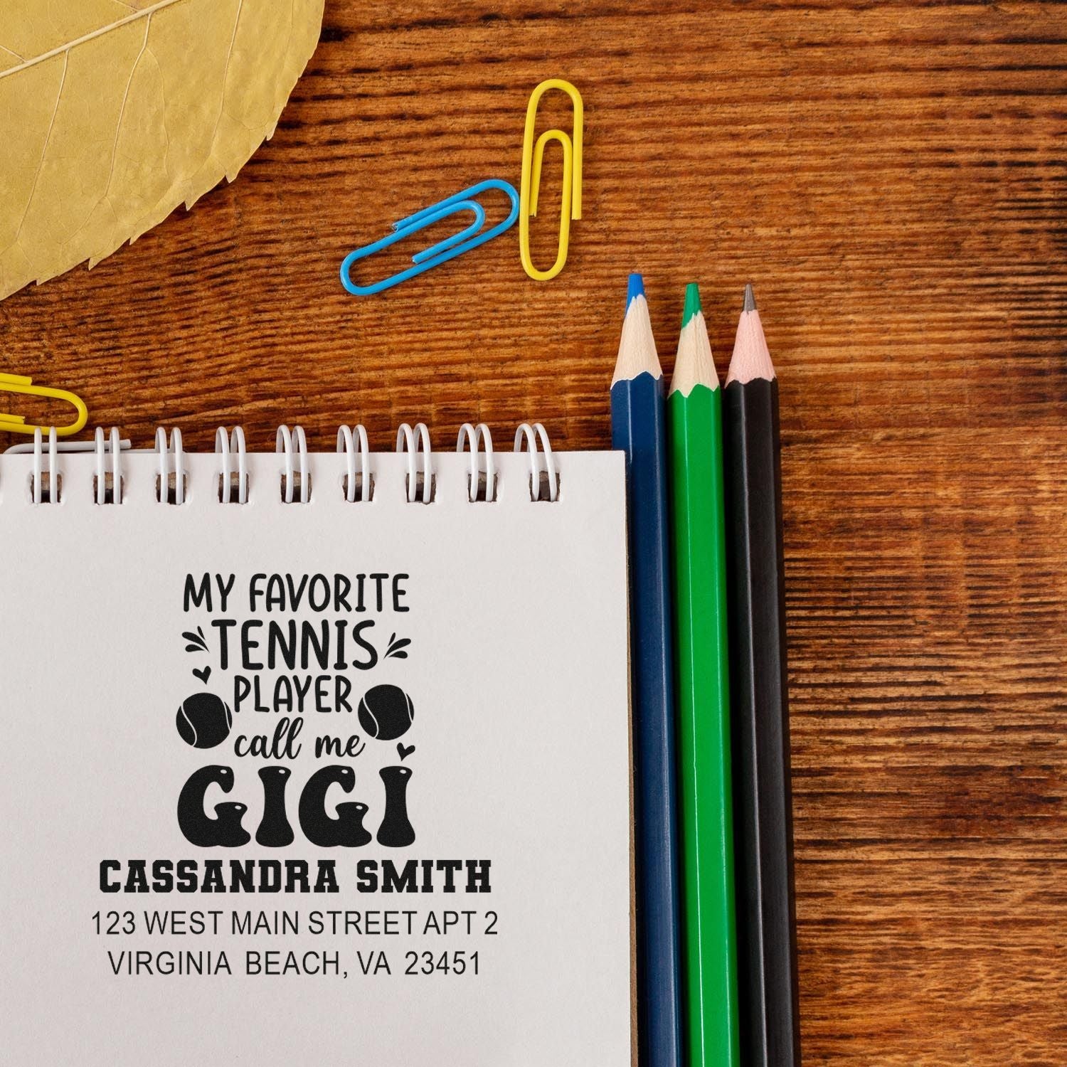 Personalized tennis rubber stamp design with custom name and address, shown in a notebook beside pencils and paper clips.