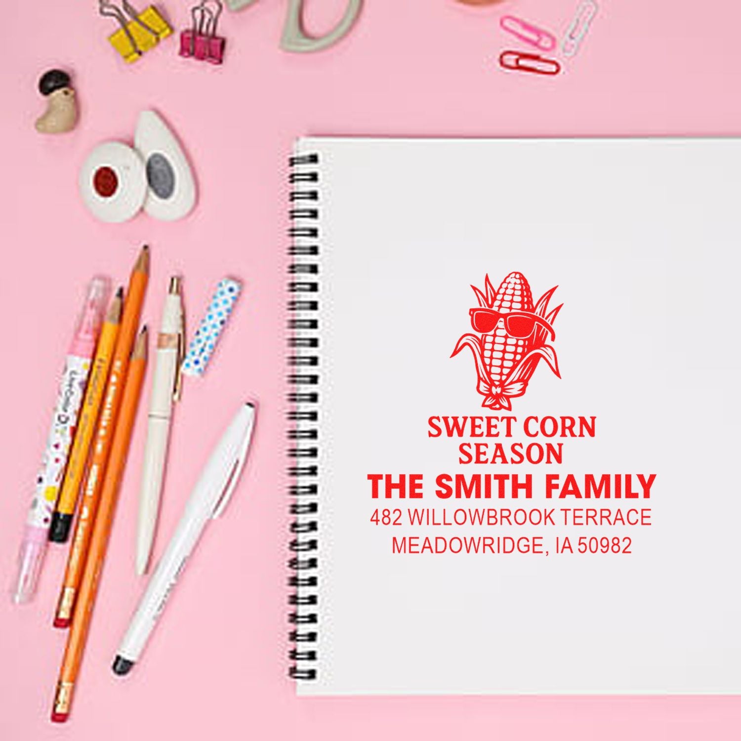 Personalized rubber stamp with sweet corn design and family address stamped on a notebook, styled with pens on a pink desk.