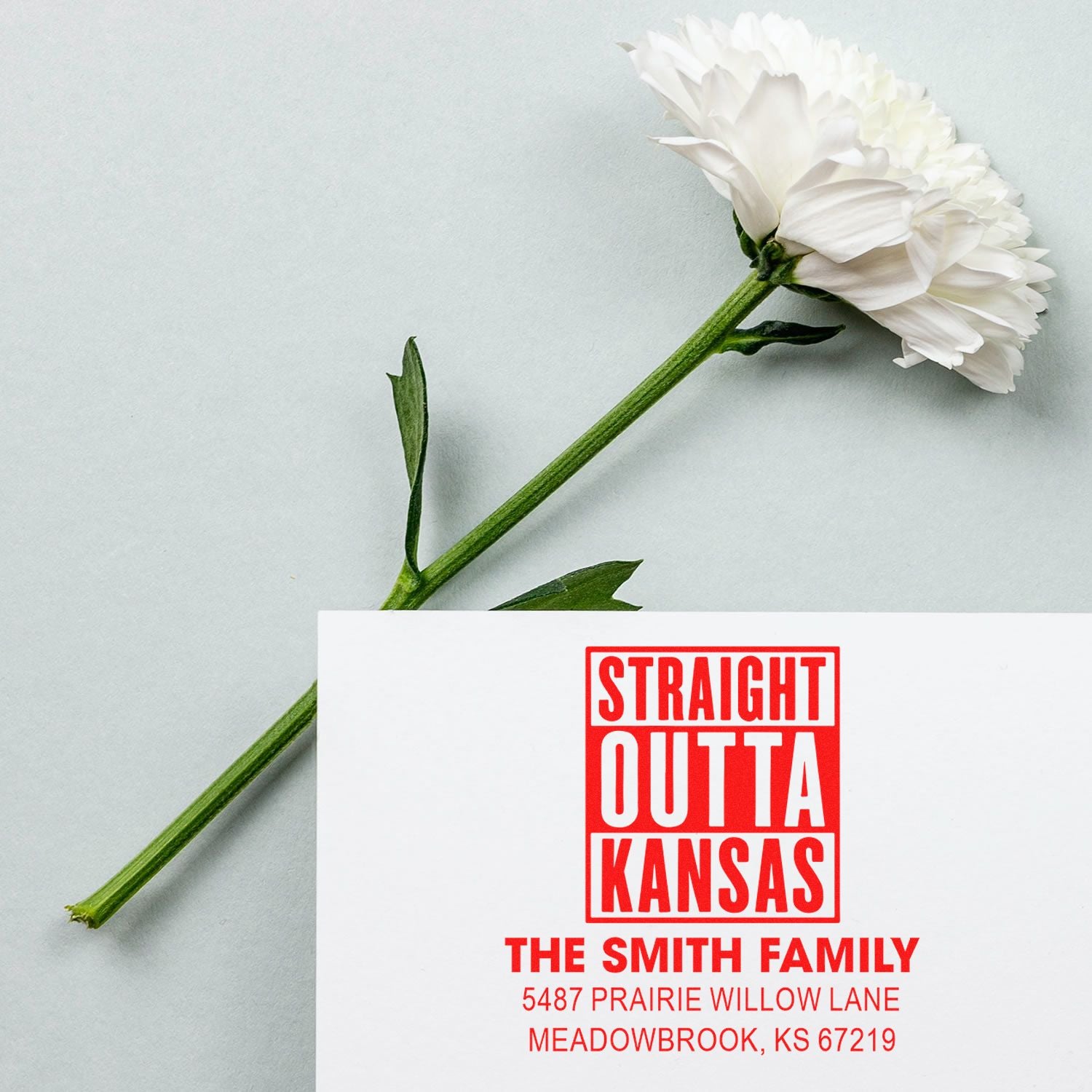 Custom rubber stamp with Straight Outta Kansas return address for The Smith Family, shown on a card beside a white flower.