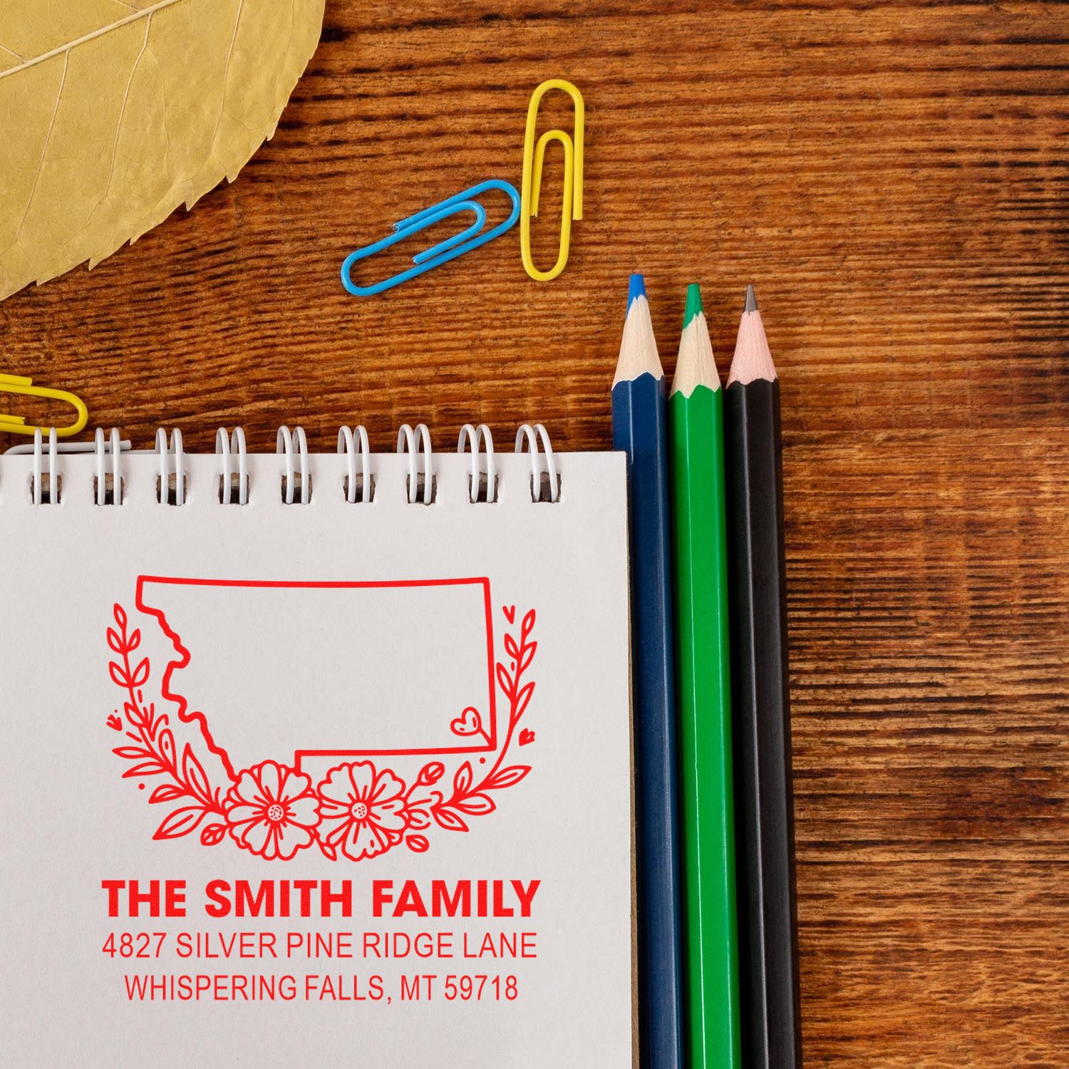 Personalized family return address rubber stamp design with floral state outline on notebook, shown with pencils and stationery on a wood desk.