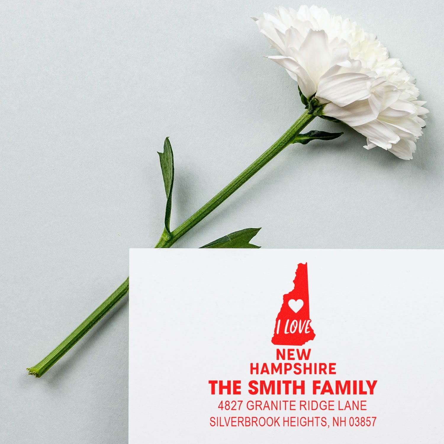 Personalized return address rubber stamp with New Hampshire design and family name on a card beside a white flower.