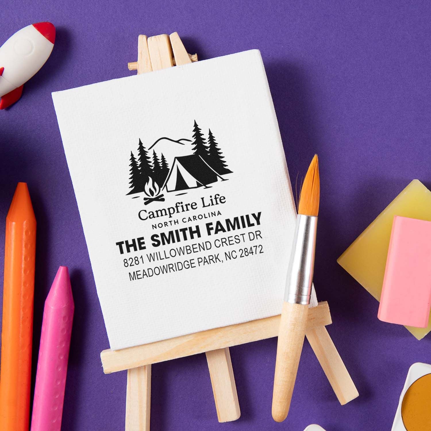 Personalized return address rubber stamp with campfire, tent and pine tree design, displayed on canvas easel.