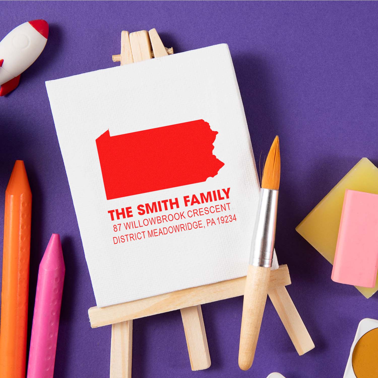 Custom family return address rubber stamp with Pennsylvania outline, displayed on an easel with art supplies.