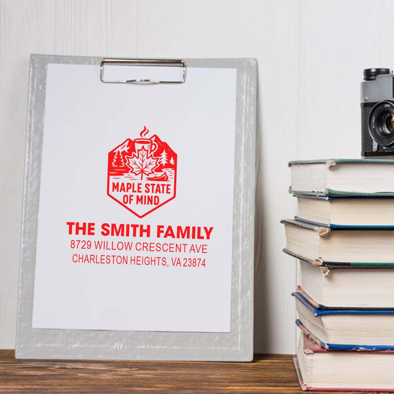 Personalized red return address stamp on clipboard, displayed on a desk beside stacked books and a camera.
