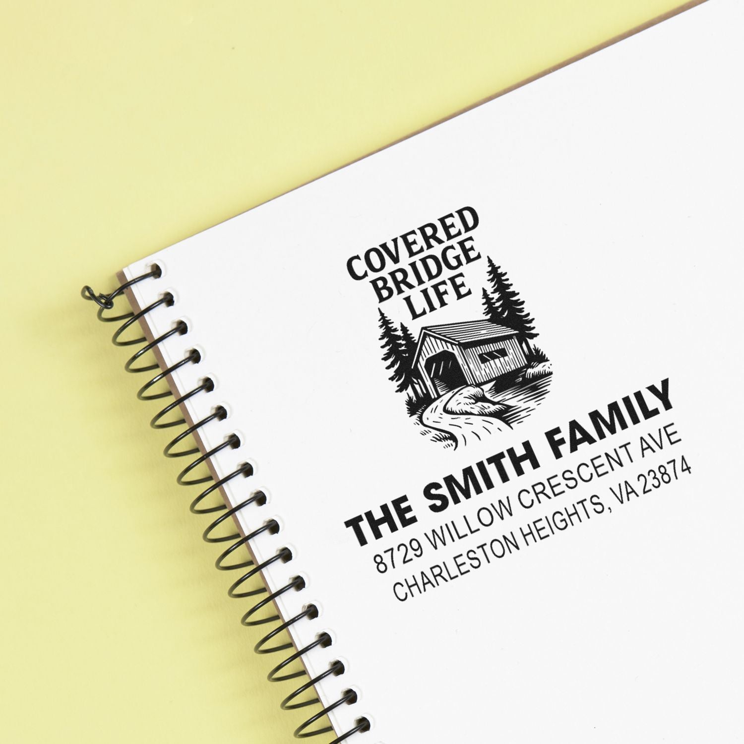 Custom address stamp with Covered Bridge Life logo, covered bridge illustration, and The Smith Family address on a spiral notebook over a yellow background.