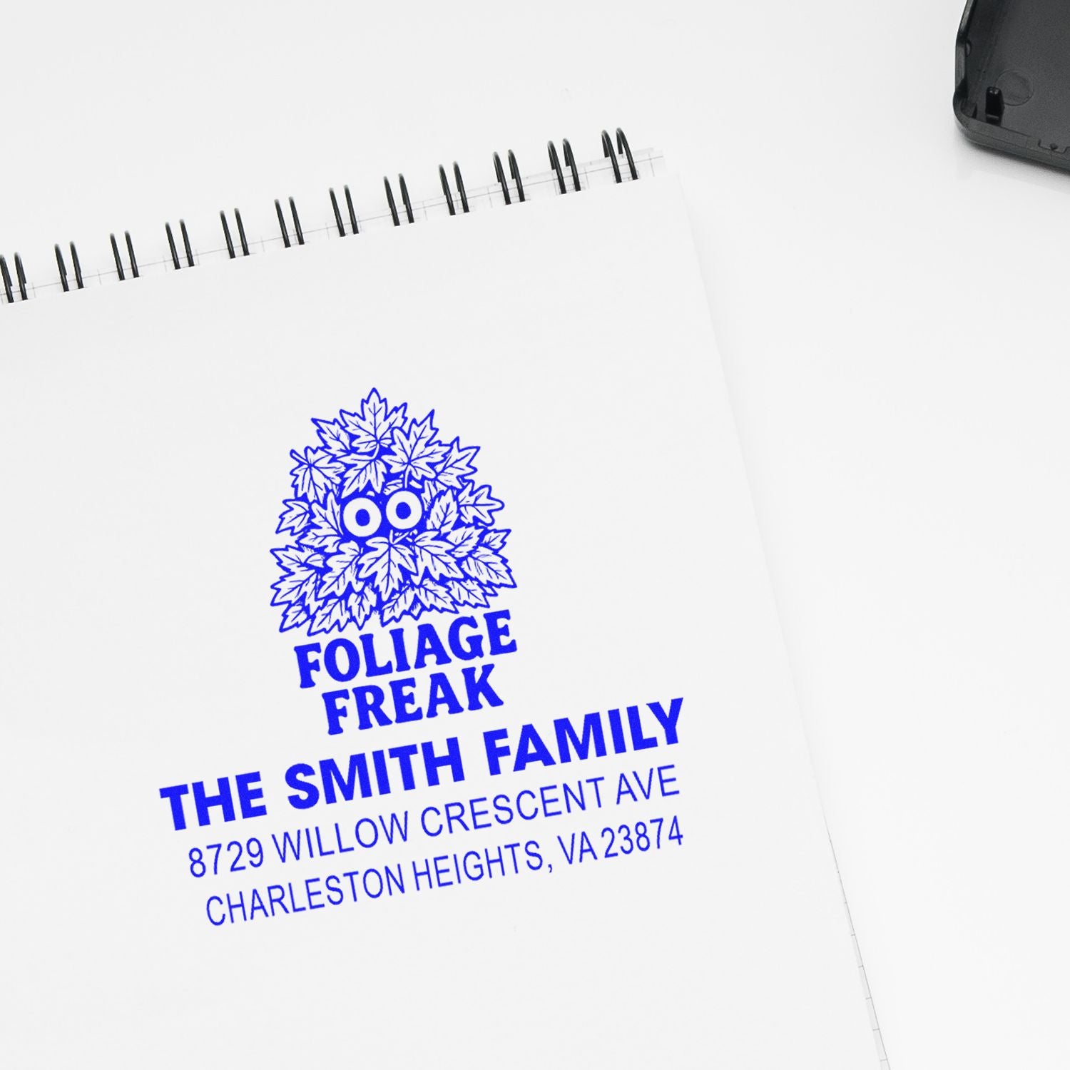 Custom blue self-inking stamp imprint on a spiral notebook, reading Foliage Freak, The Smith Family with address details on a white desk.