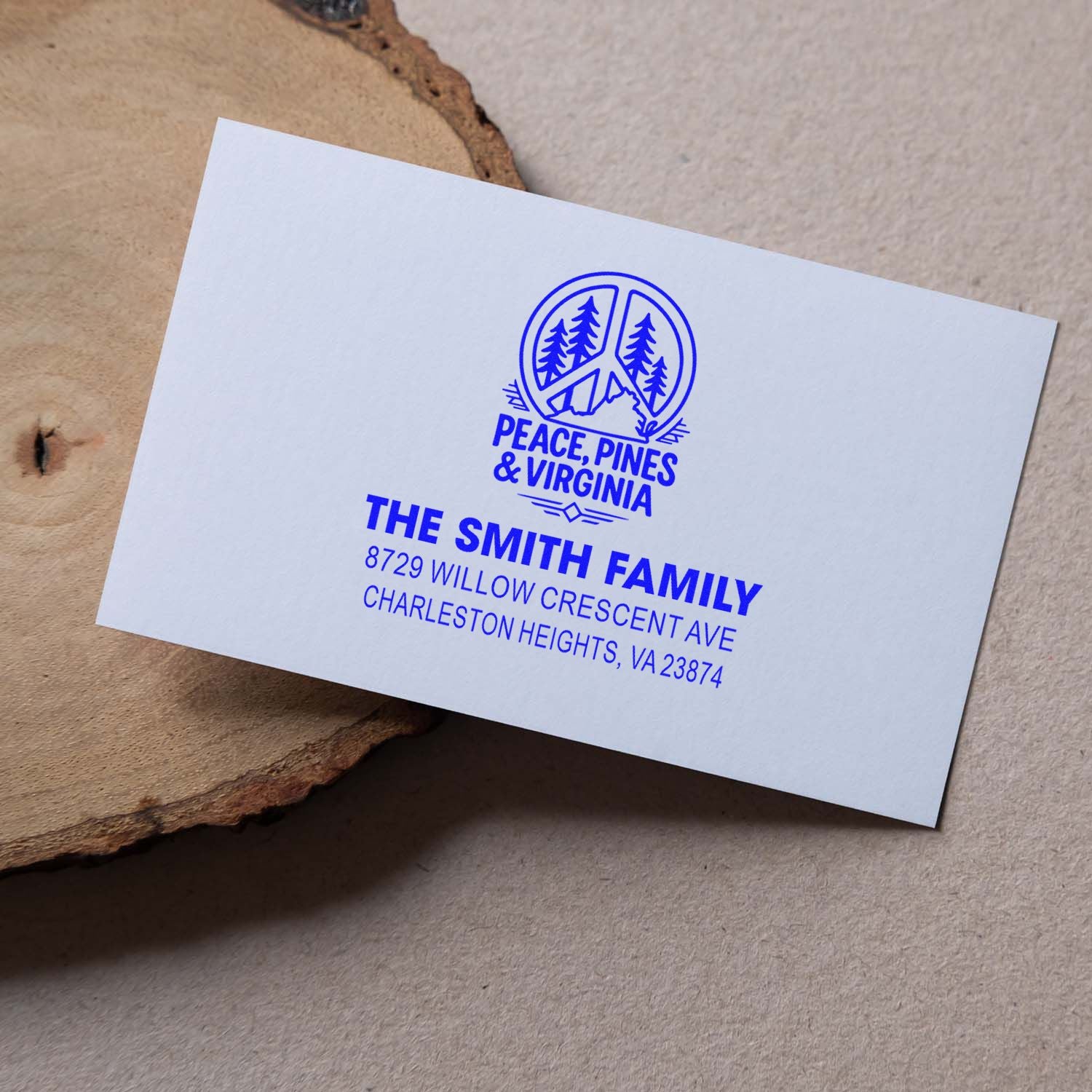 Blue custom return address stamp on a white card, featuring Peace, Pines & Virginia and The Smith Family, on a rustic wood slice.