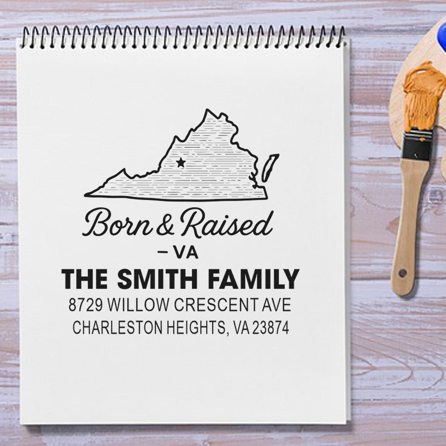 Custom Virginia return address stamp on a spiral notebook, with paintbrush on a wood table, featuring a state outline and The Smith Family address.