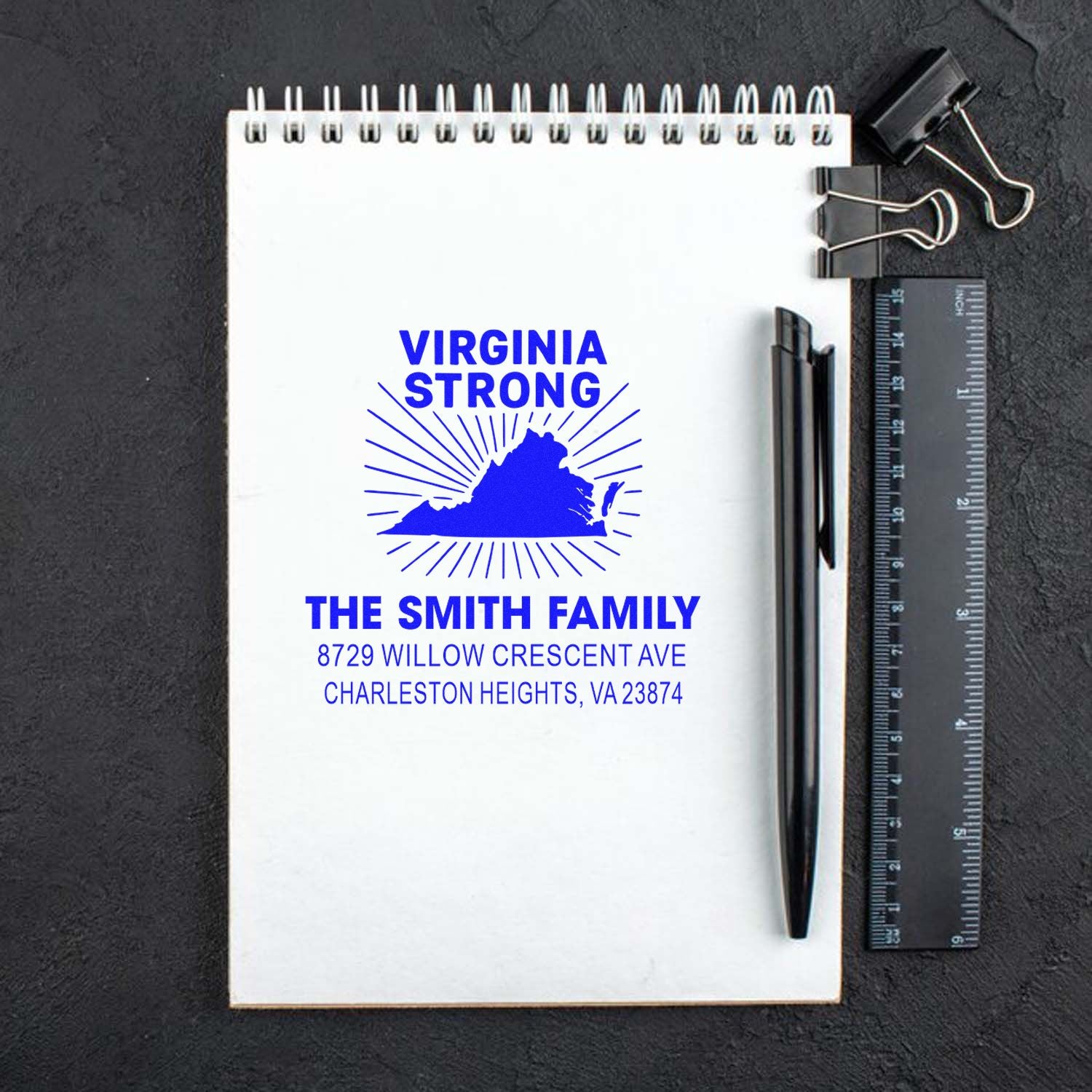 Custom Virginia Strong return address stamp on white notepad, with pen, ruler and binder clips on a dark desk background.