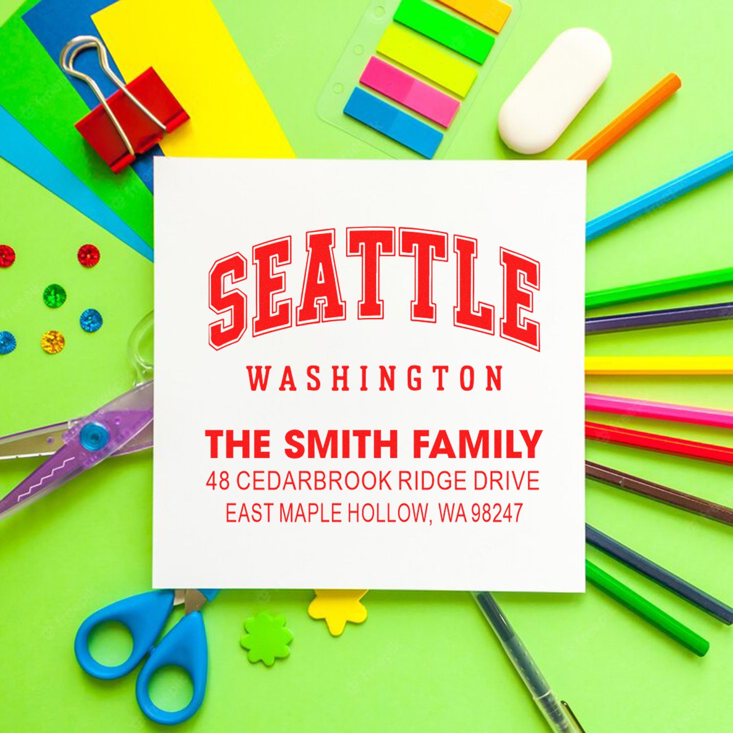 Custom Seattle Washington address stamp for The Smith Family, shown with colorful stationery, binder clips, scissors, and pencils on a green background.