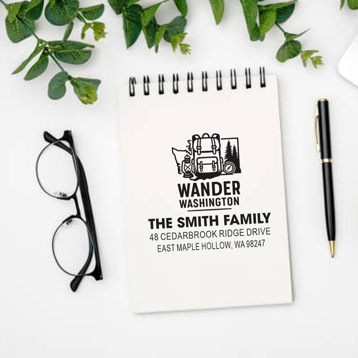 Custom family address stamp on spiral notebook with WANDER WASHINGTON travel design, styled on a white desk with glasses, pen, and greenery.