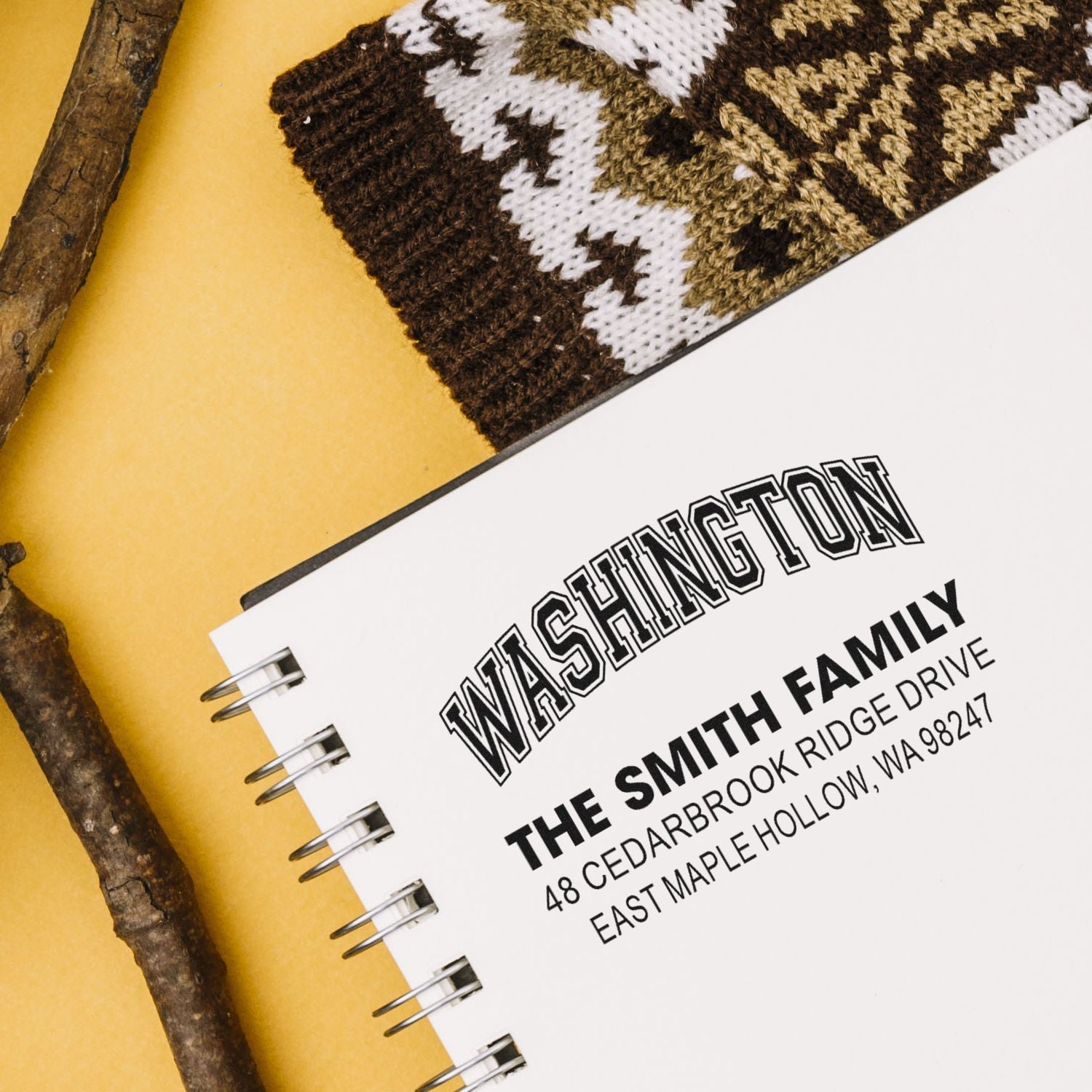 Custom Washington family address stamp on a spiral notebook, styled with woven textile and rustic branches on a yellow background.