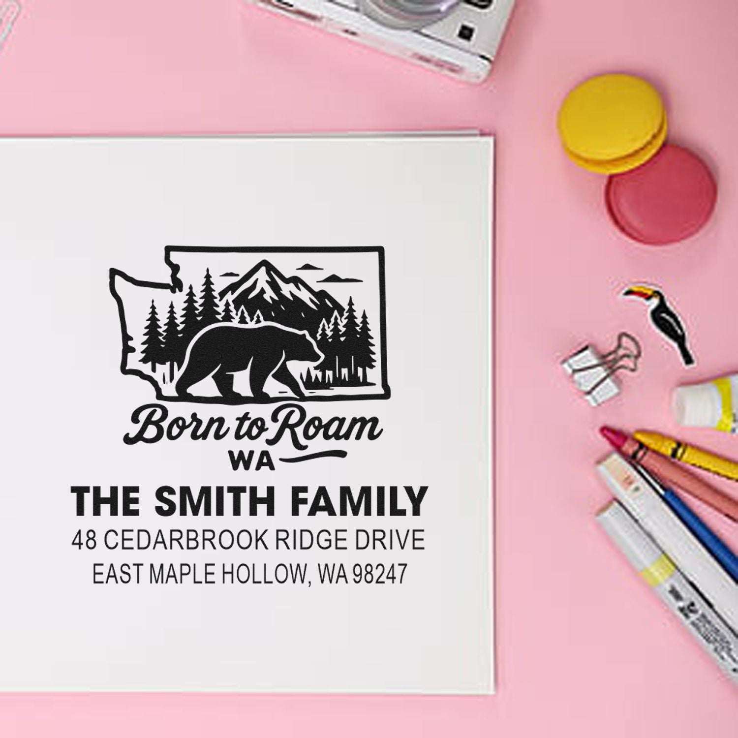 Personalized rubber address stamp for The Smith Family with Washington bear and mountain design on white paper, on a pink desk with ink pads and markers.