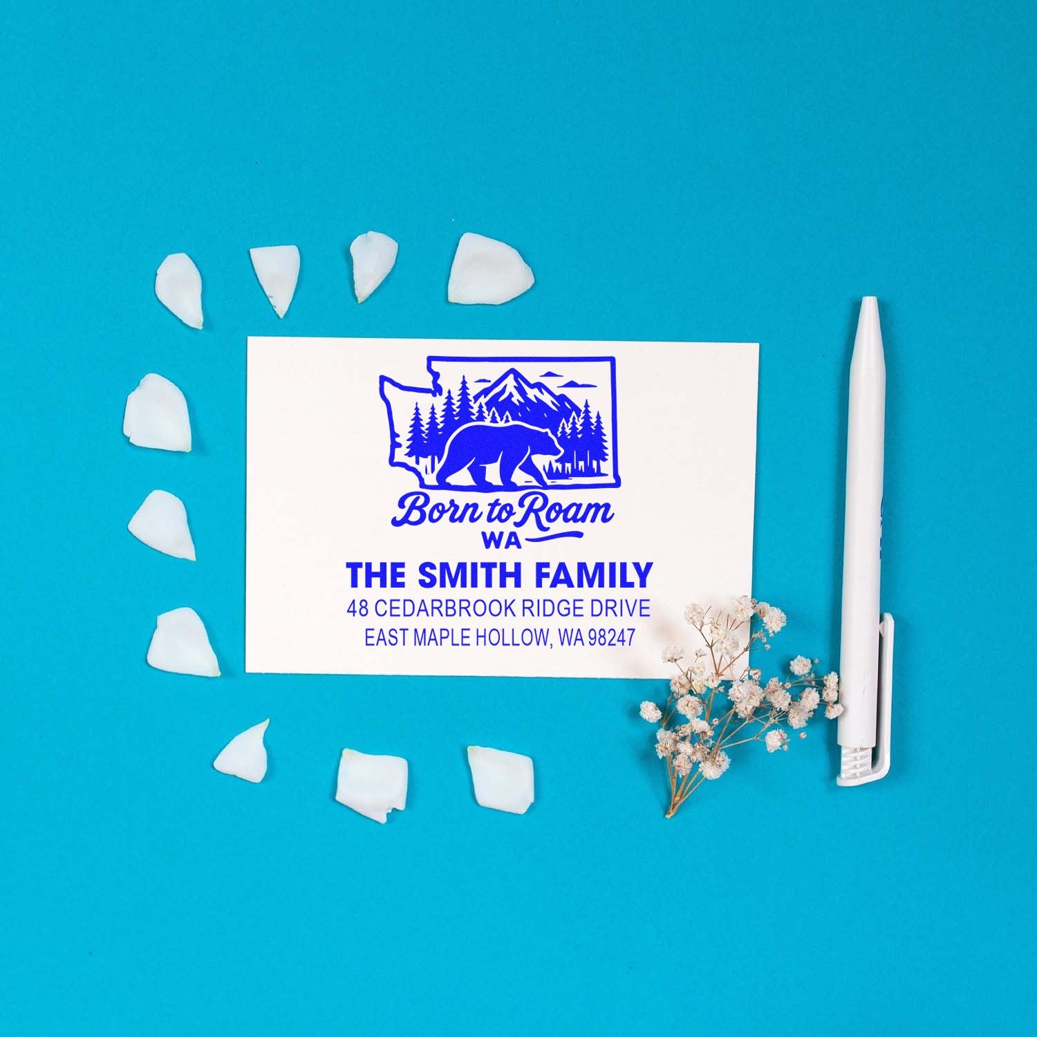 Personalized family address stamp with bear and mountain design on blue background, styled with white pen, petals, and dried flowers.