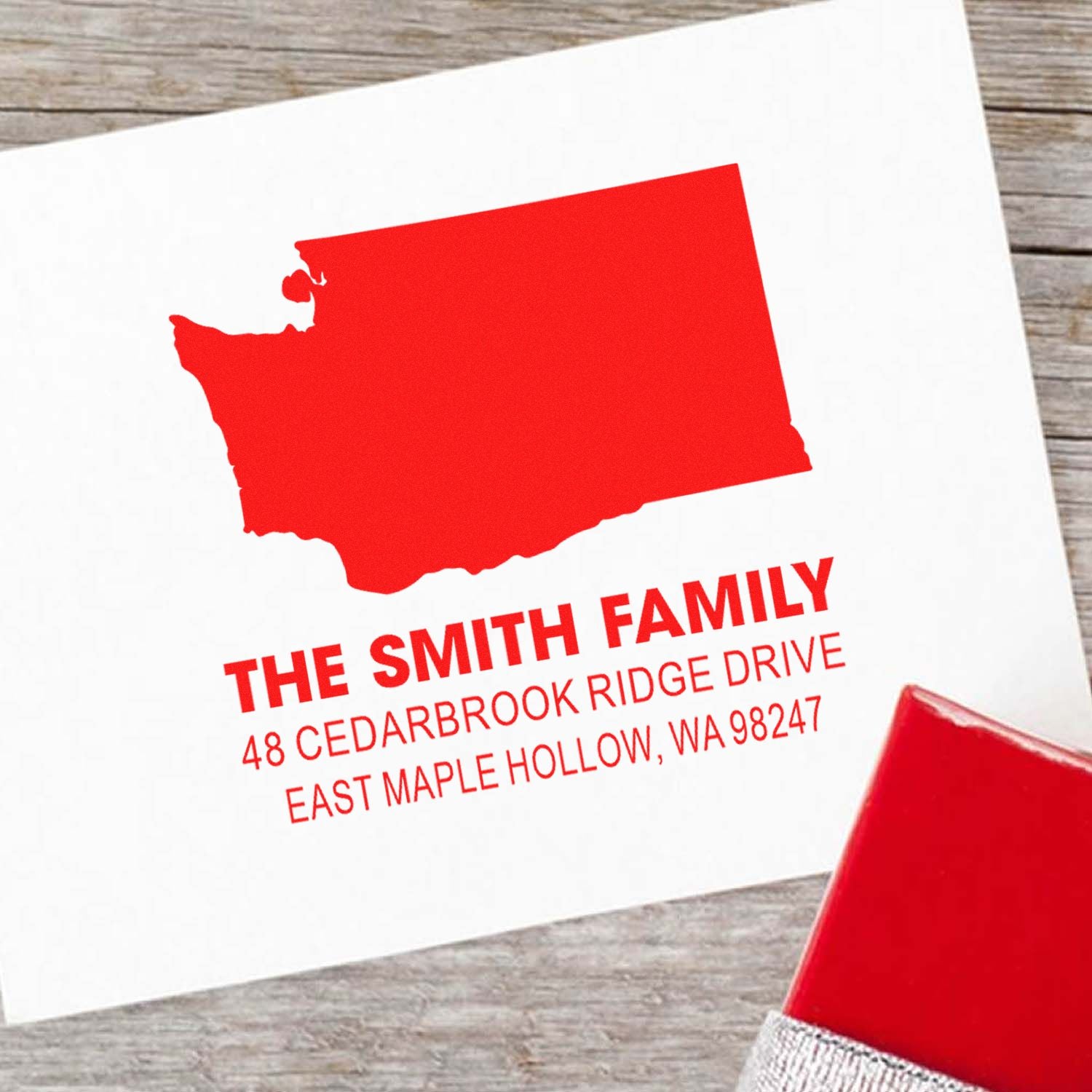 Custom red family return address stamp with Washington state silhouette on white paper, displayed on a wooden table with stamp handle visible.