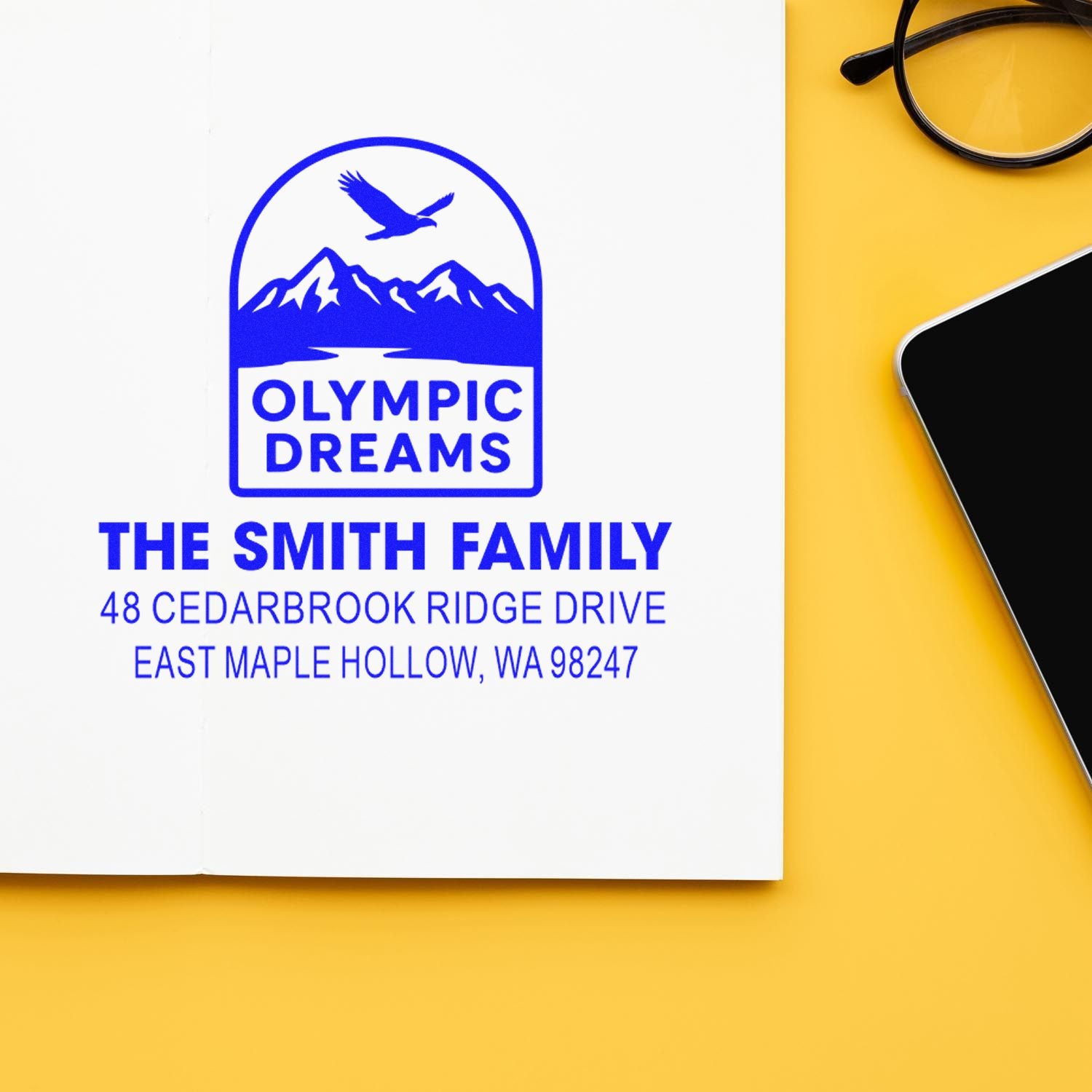 Custom family address stamp with Olympic Dreams mountain and eagle logo on white paper, beside glasses and phone on yellow desk.