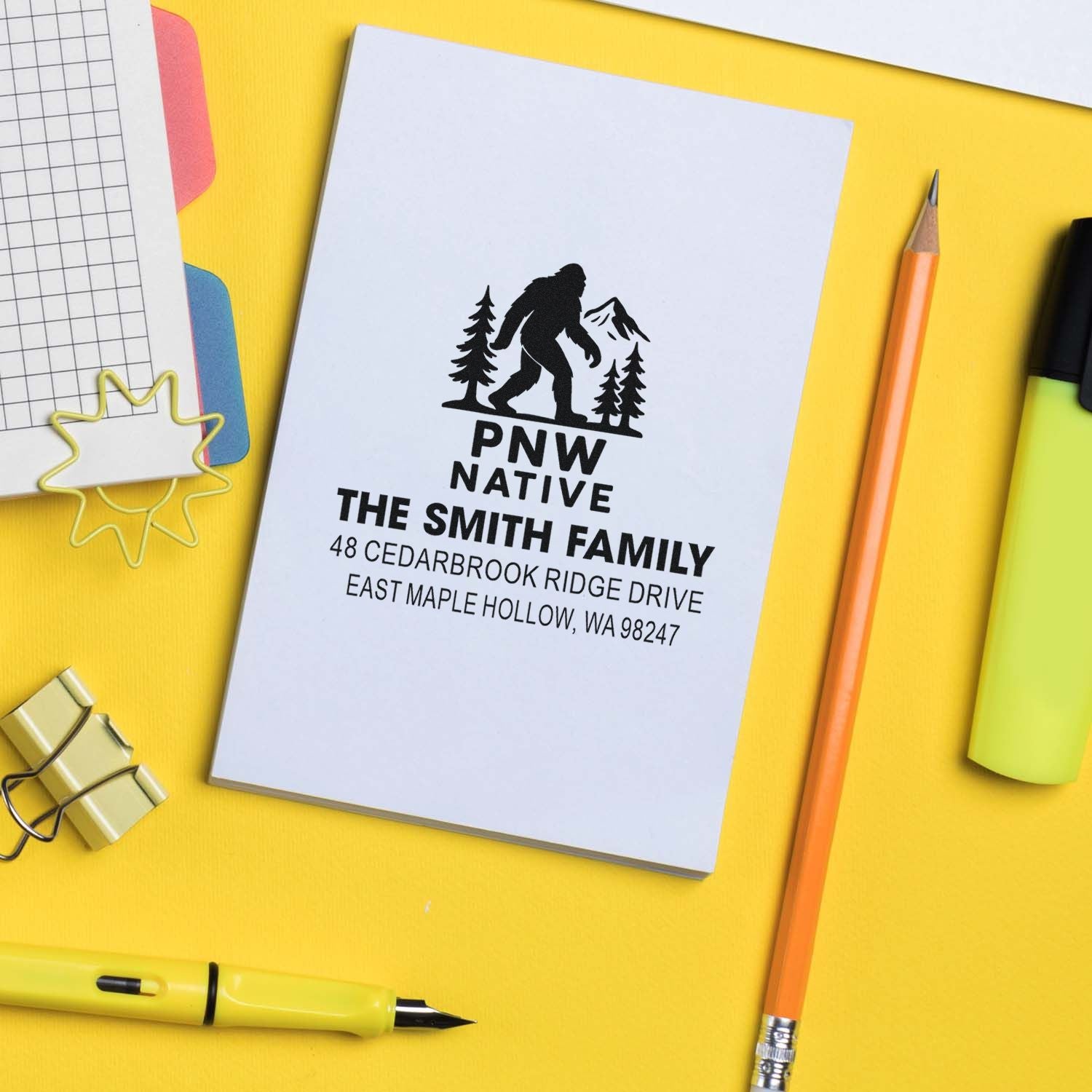 Custom PNW Native family address stamp with Bigfoot, pine trees and mountains on a notebook, surrounded by pencil, pen and stationery.