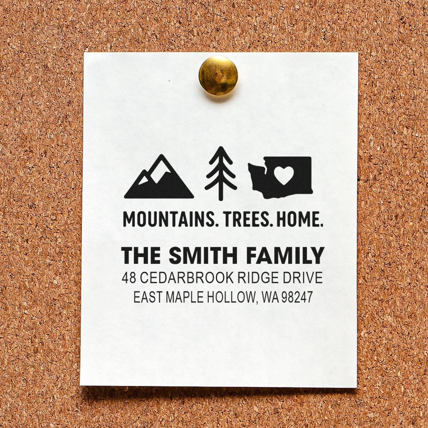Custom address stamp on a white card pinned to a corkboard, featuring mountains, trees, home icons, and The Smith Family address.