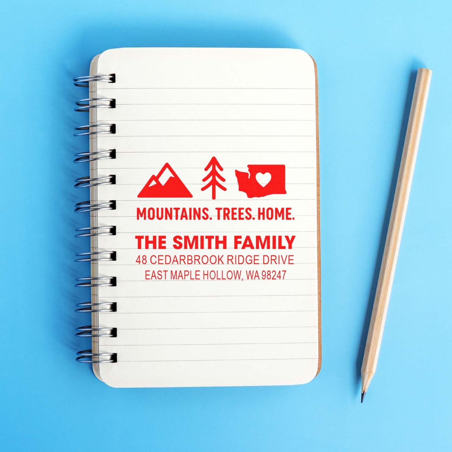 Custom address stamp on a spiral notebook with red mountains, trees and heart state design, beside a pencil on a blue background.