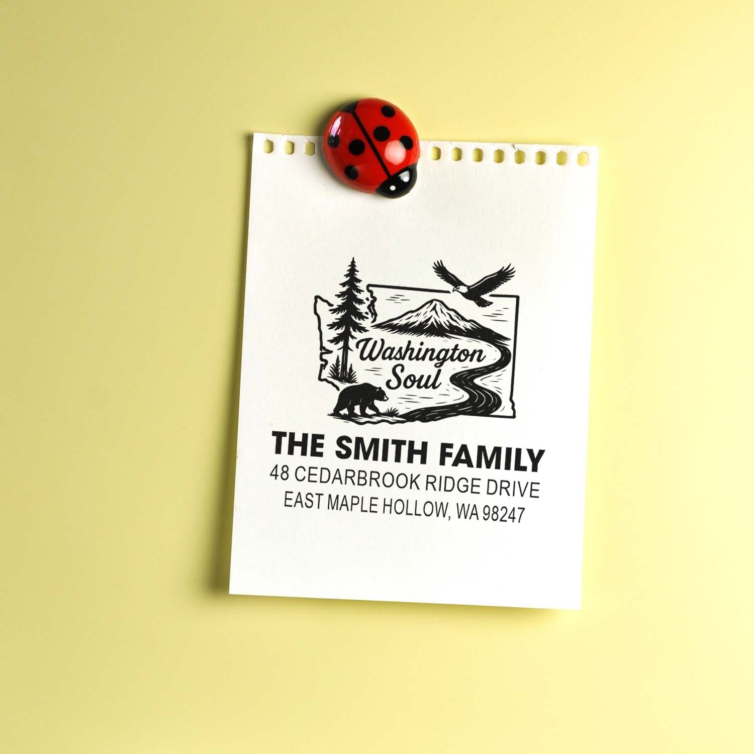 Custom Washington family address stamp with mountain, eagle and bear design, clipped to paper by a ladybug on a yellow background.