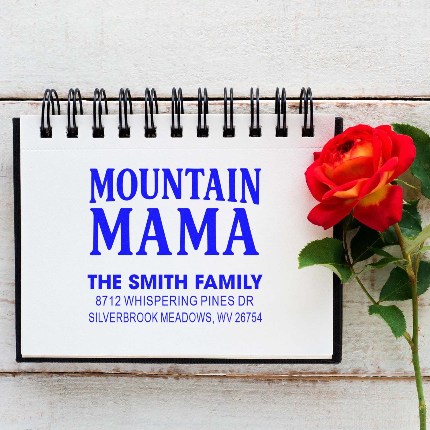 Custom address stamp for Mountain Mama, The Smith Family, shown on a spiral notebook beside a red rose on white wood.