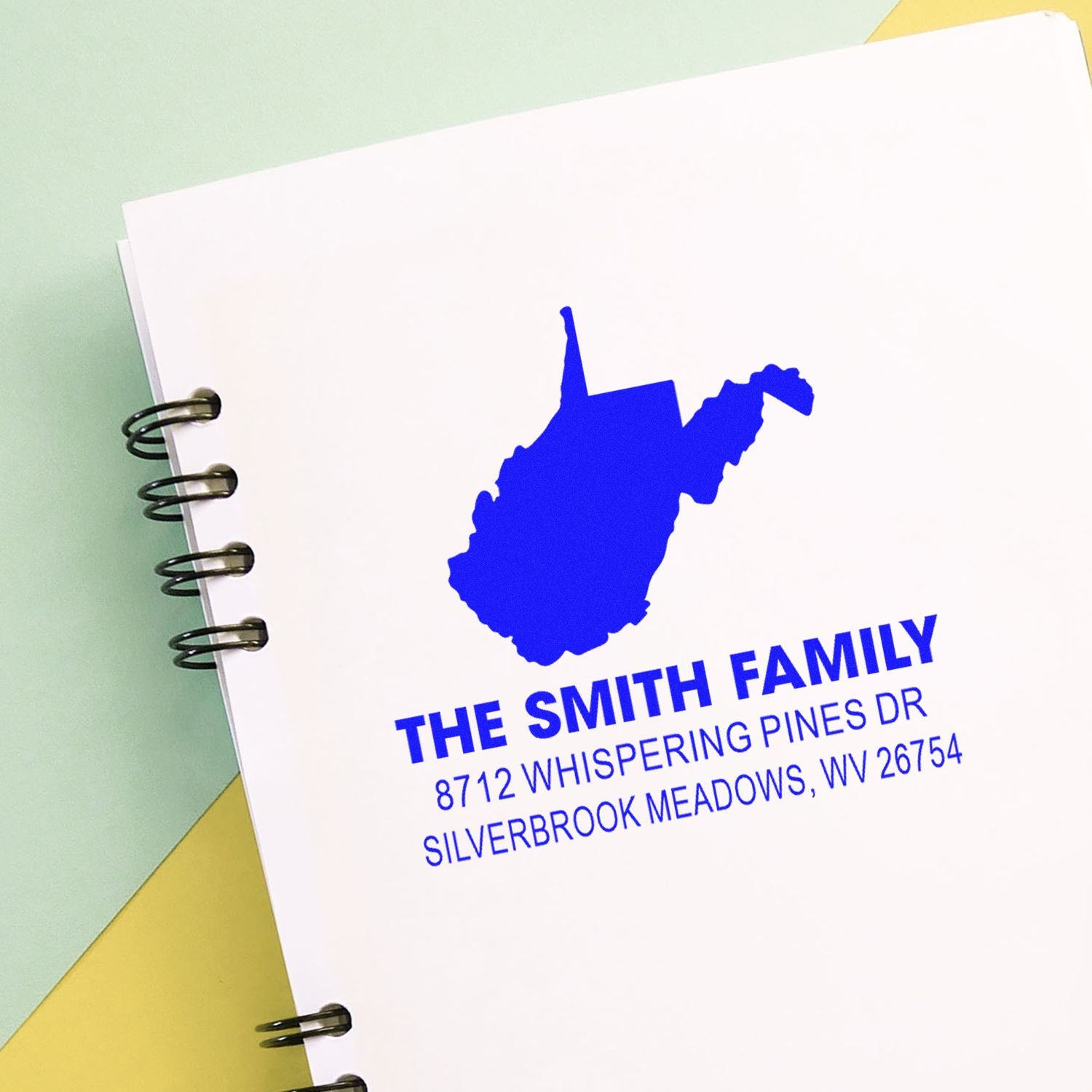 Custom blue self-inking address stamp with West Virginia silhouette on a spiral notebook, reading The Smith Family, 8712 Whispering Pines Dr, Silverbrook Meadows, WV.