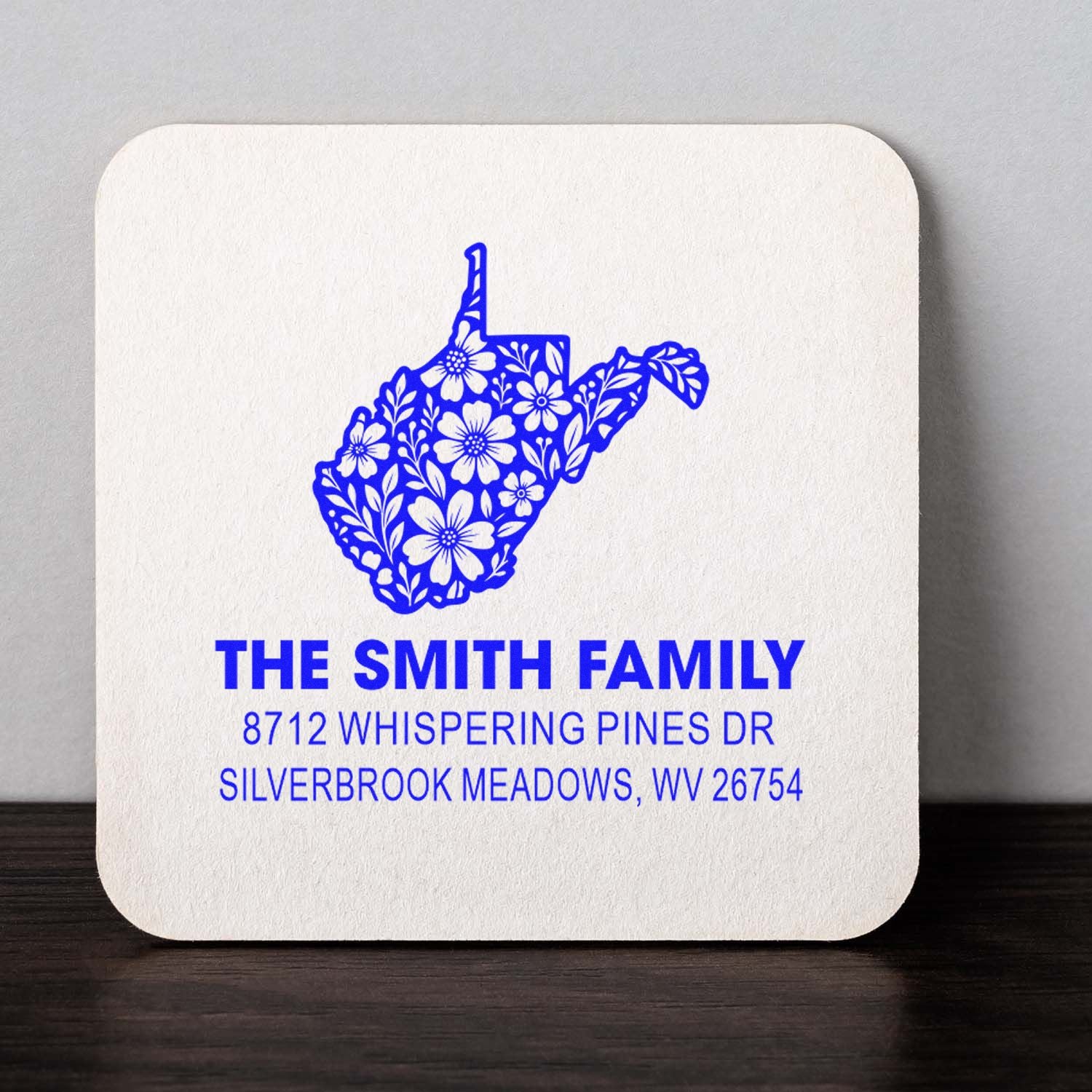 Custom address stamp with floral West Virginia silhouette and Smith Family address in blue ink on white cardstock over a dark tabletop.