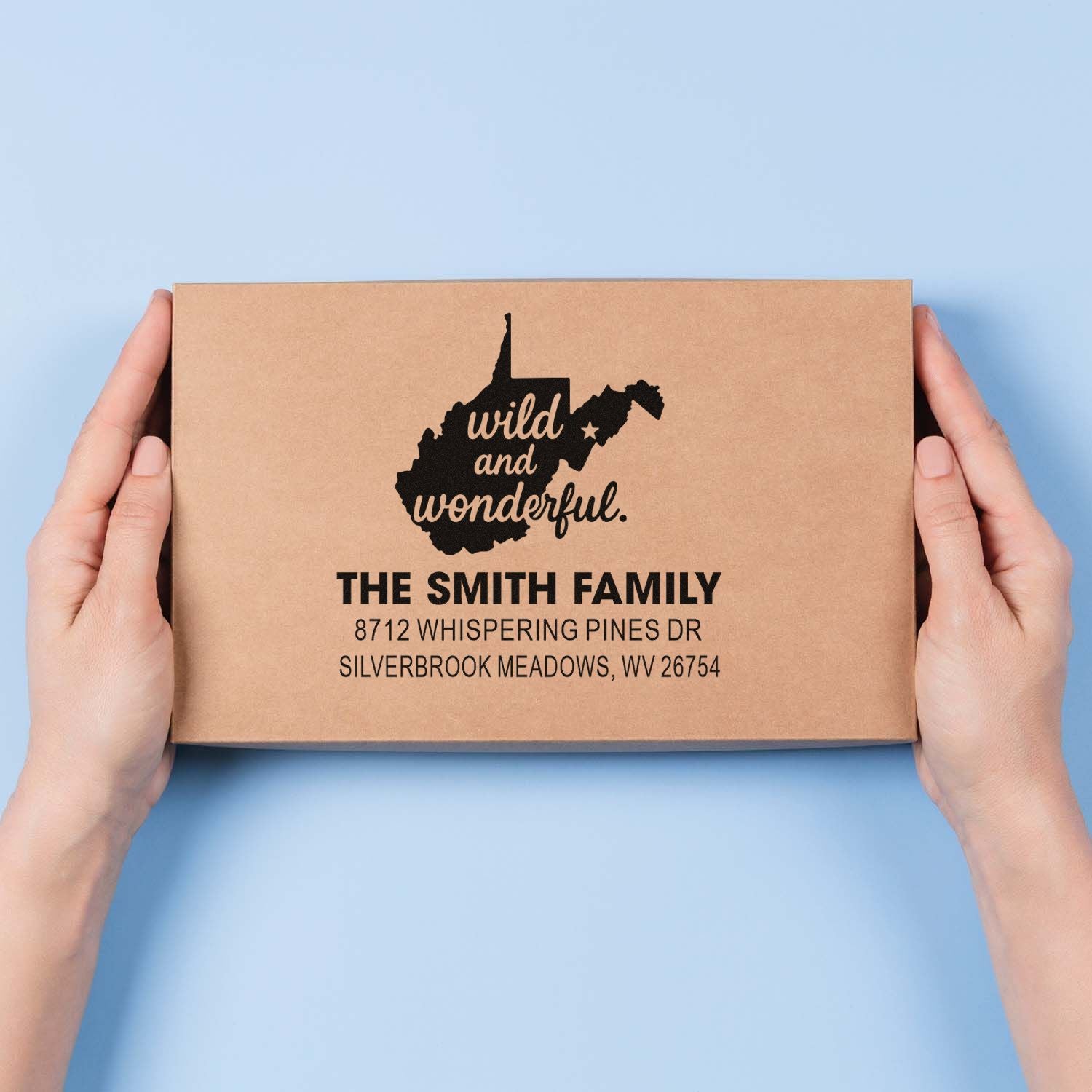 Hands holding kraft mailer stamped with West Virginia silhouette and Wild and Wonderful custom family address stamp on a blue background.