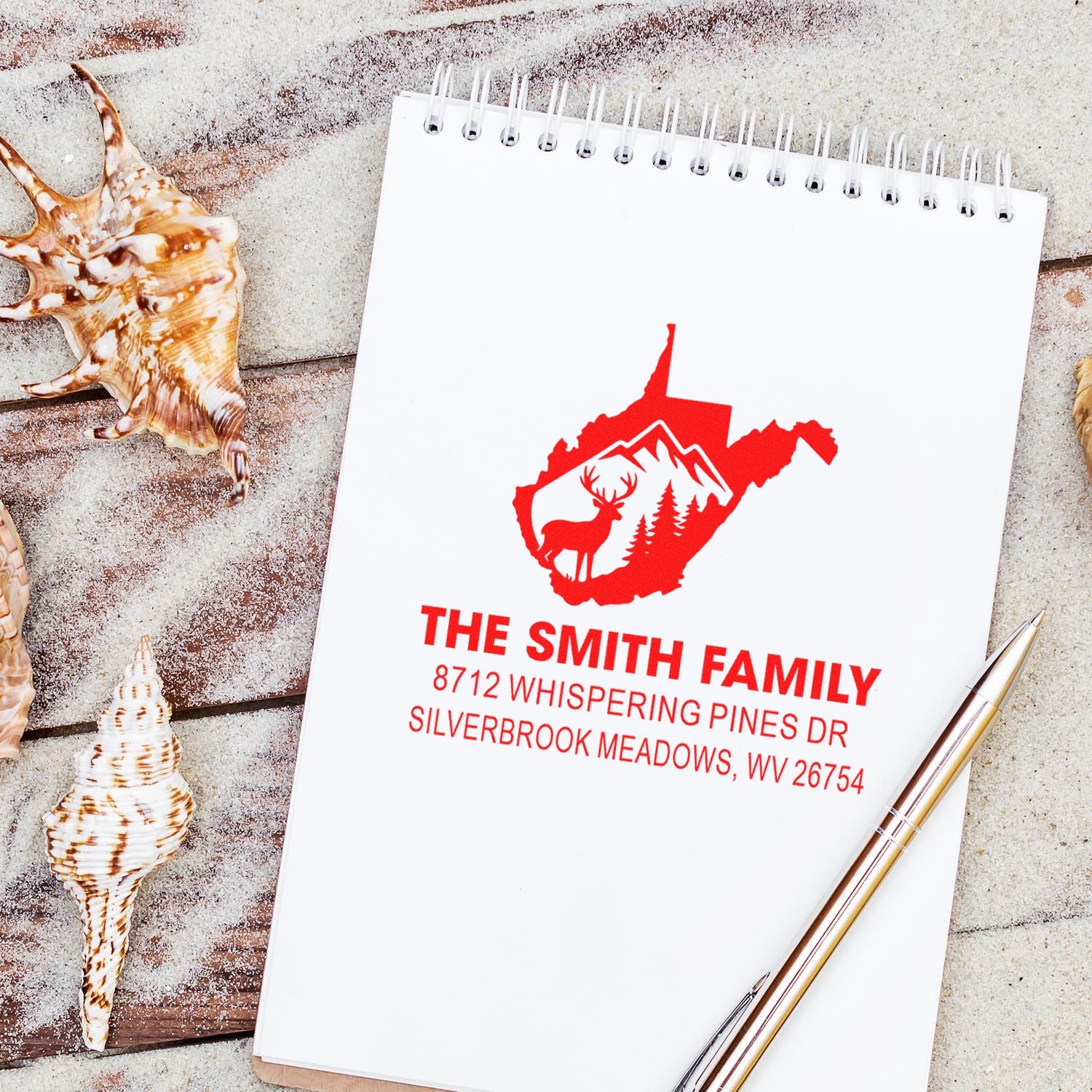 Custom family return address stamp on spiral notepad with pen and seashells, featuring deer and mountain logo on a beach-style rustic background.