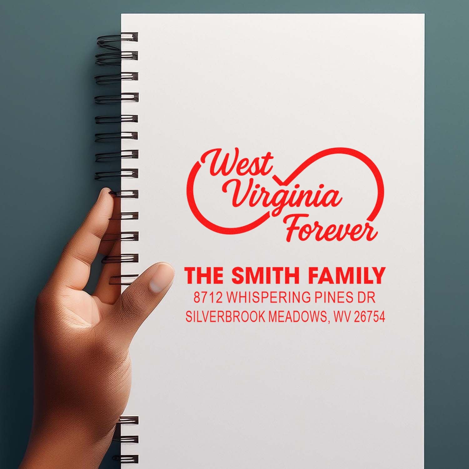 Hand holding a spiral notebook featuring a red West Virginia Forever custom family address stamp for The Smith Family on a white cover.