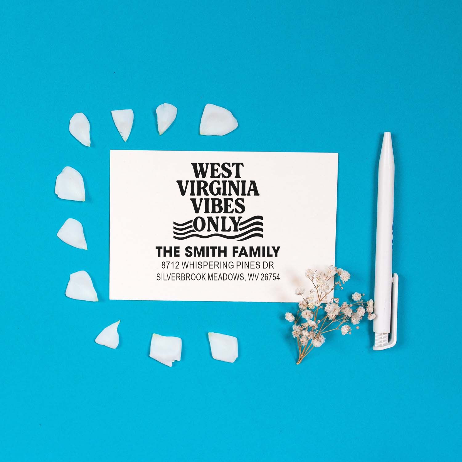 Custom address stamp reading West Virginia Vibes Only for The Smith Family, styled on a blue background with white petals and a stamp pen.
