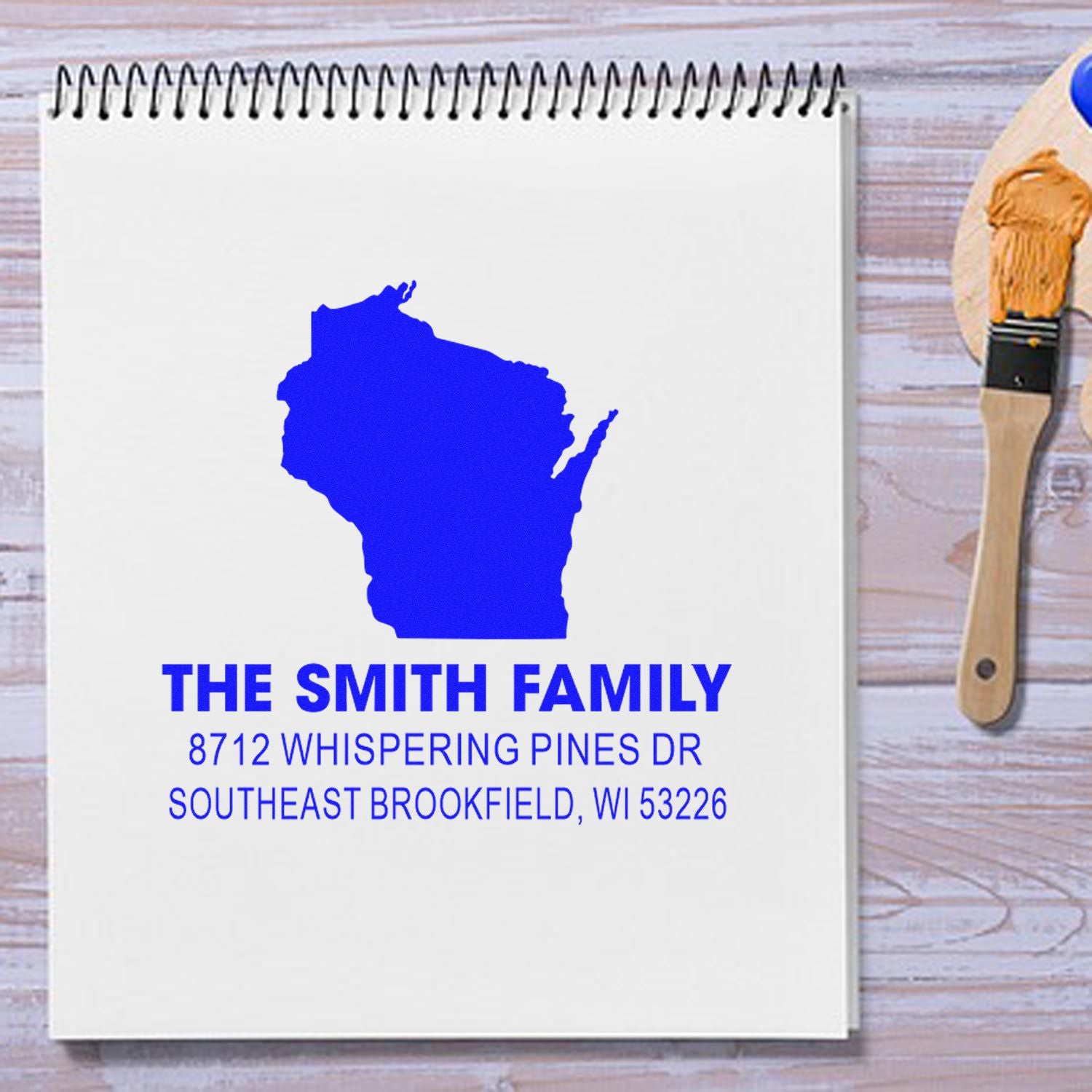 Personalized family address stamp with Wisconsin silhouette on spiral notebook, beside paintbrush on rustic wood desk.
