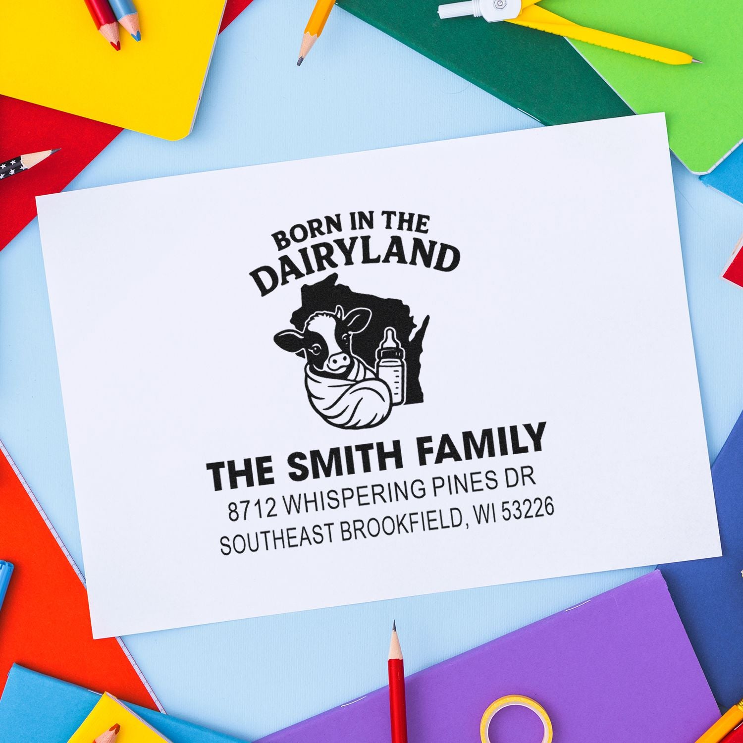 Custom Born in the Dairyland address stamp for The Smith Family, with cow and baby bottle, on white paper surrounded by colorful pencils.