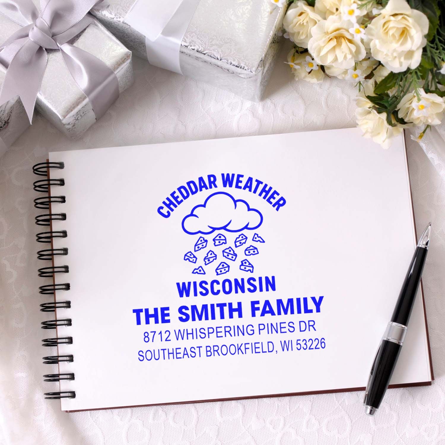 Custom Cheddar Weather Wisconsin family address stamp on a spiral notebook, with gift boxes, flowers, and a pen on a white table.